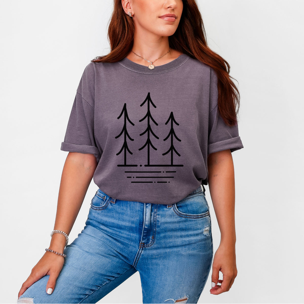 Woman wearing a Comfort Colors t-shirt printed with black line art of three simple pine trees above horizontal lines.