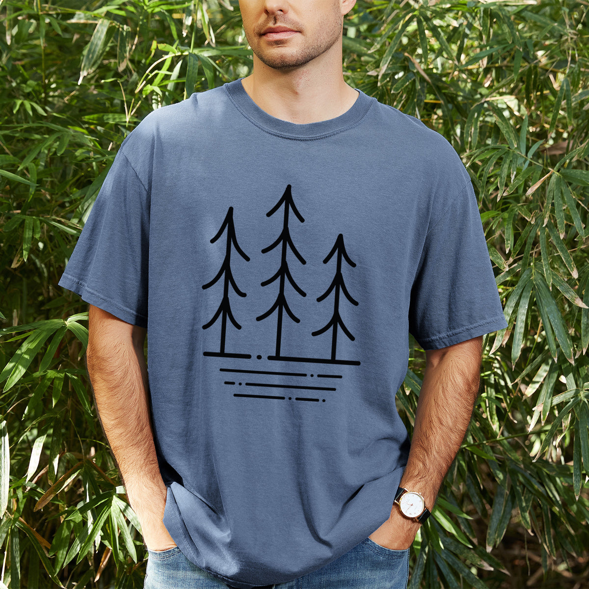 Man wearing a Comfort Colors t-shirt printed with black line art of three simple pine trees above horizontal lines, outdoors.