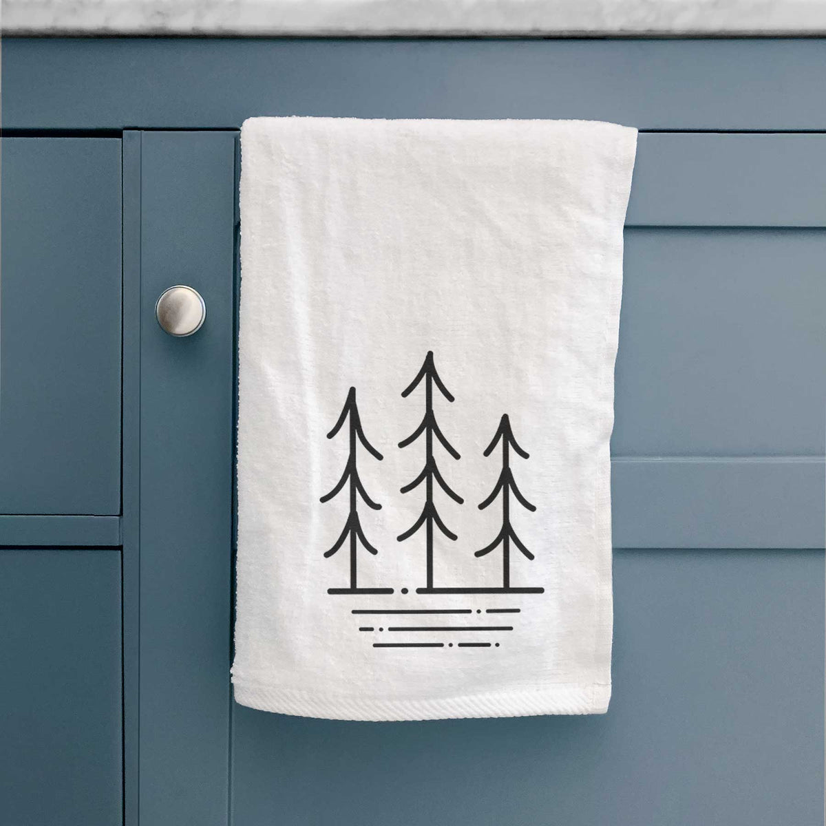 White hand towel printed with black line art of three simple pine trees above horizontal lines, hanging on a cabinet door.