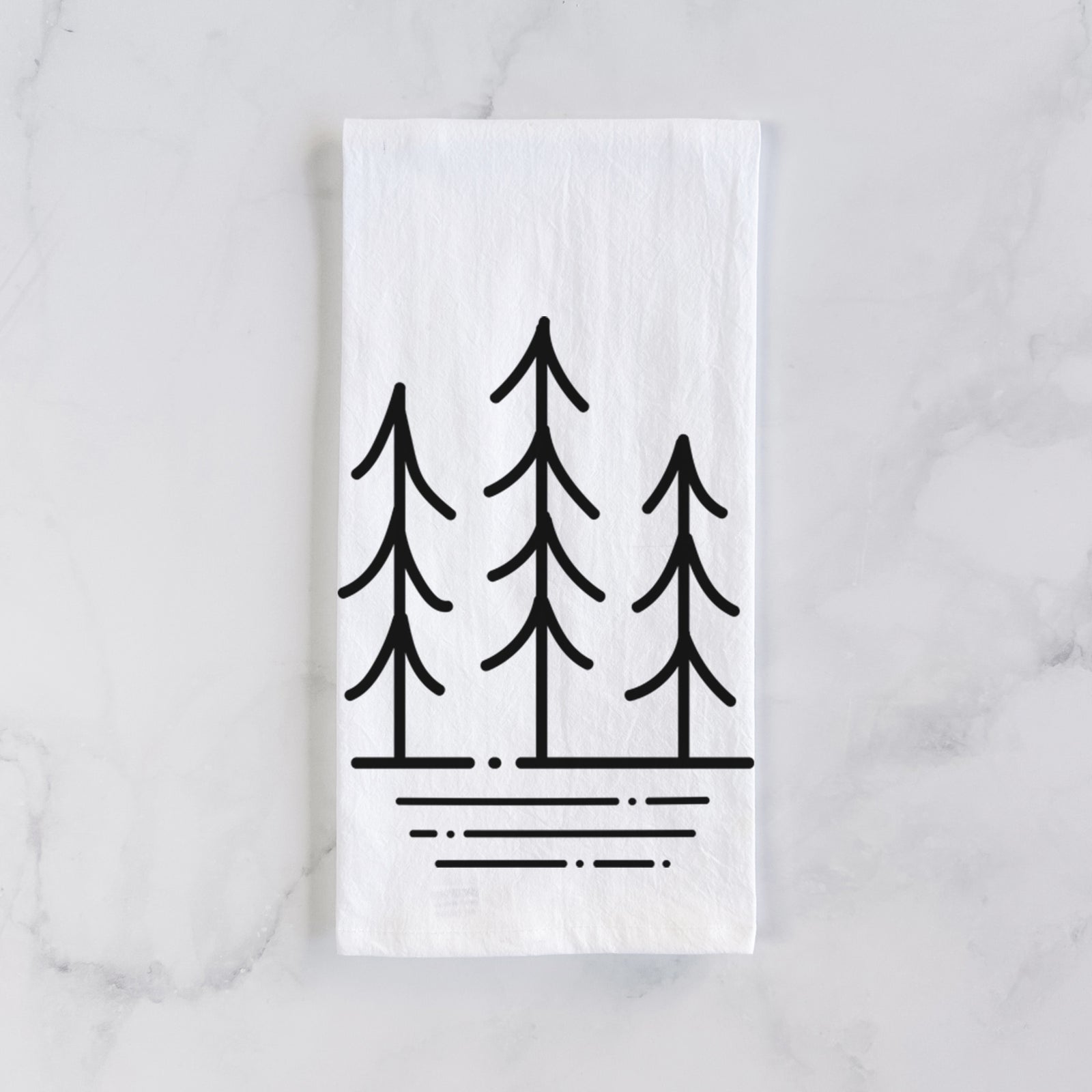 White cotton tea towel printed with black line art of three simple pine trees above horizontal lines, on a marble counter beside a round wooden cutting board and greenery.