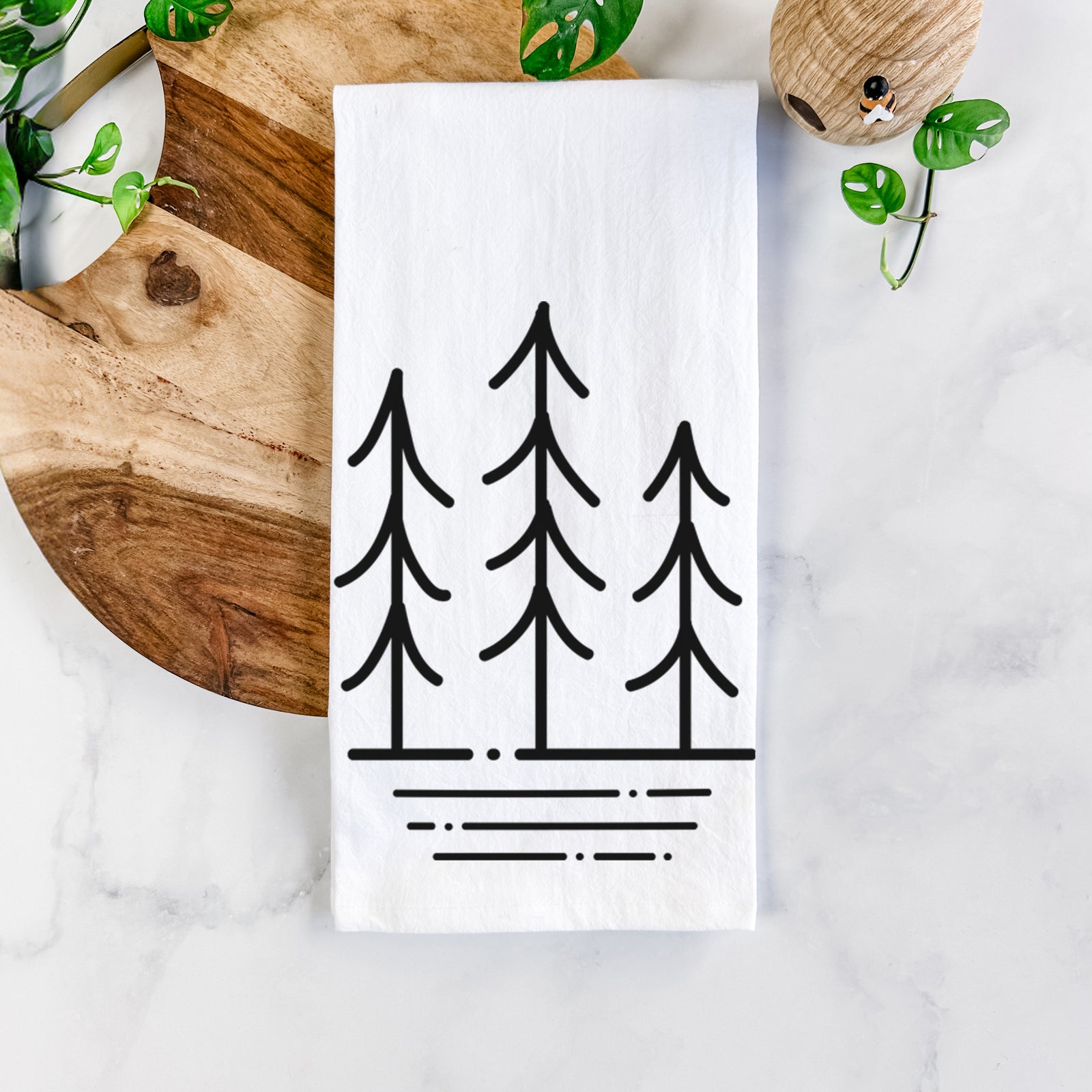 White cotton tea towel printed with black line art of three simple pine trees above horizontal lines, on a marble counter beside a round wooden cutting board and greenery.
