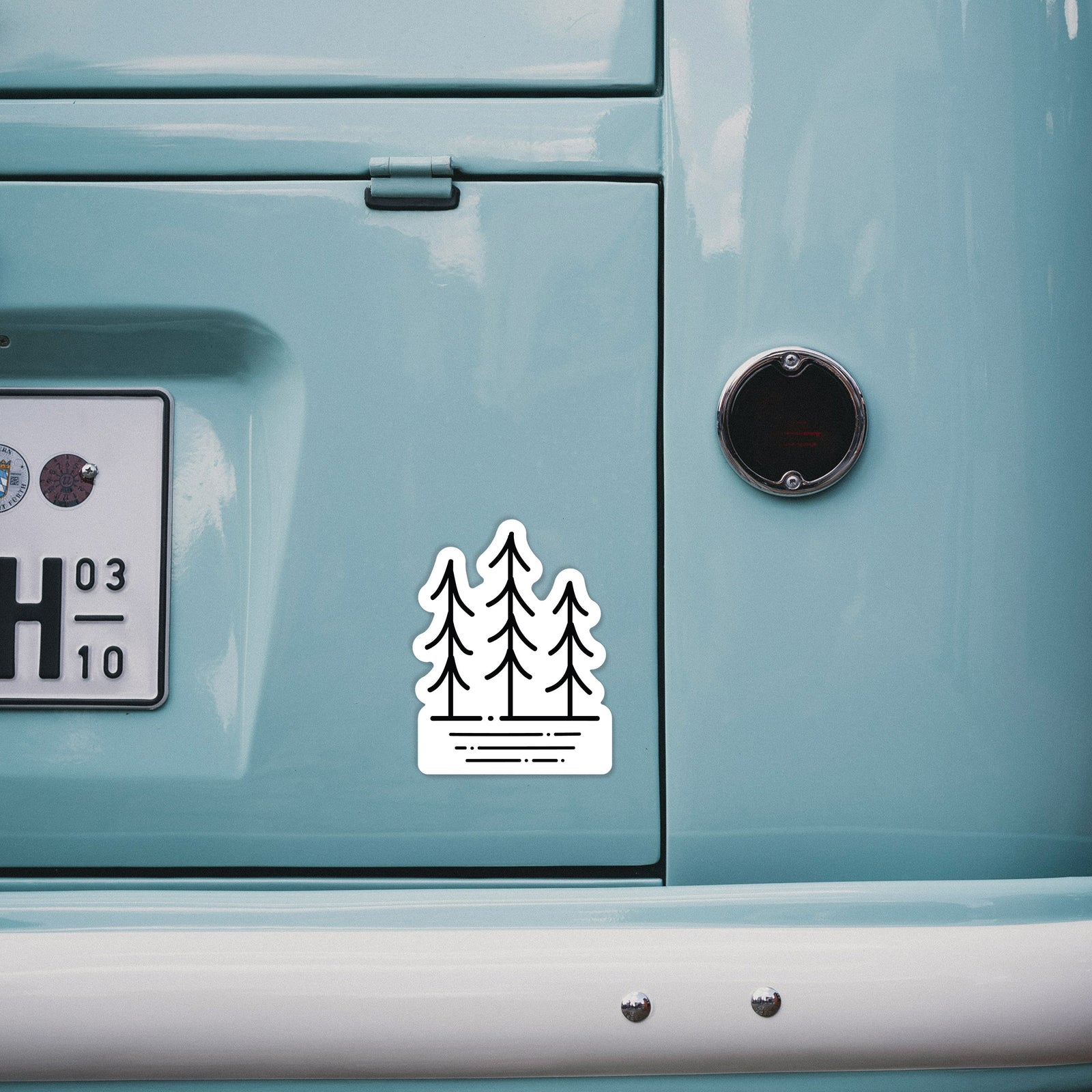 Hand holding a 4 inch vinyl decal of black line art of three simple pine trees above horizontal lines.