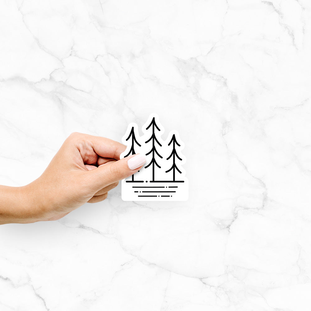 Hand holding a 4 inch vinyl decal of black line art of three simple pine trees above horizontal lines.