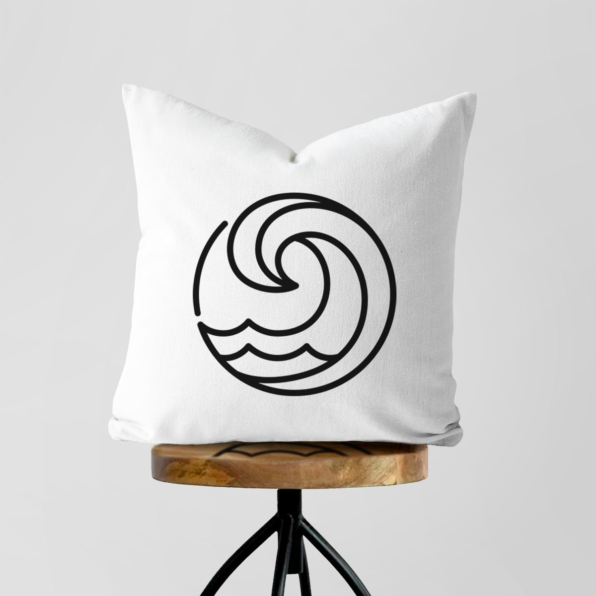 White pillow cover printed with a bold black line art circle of a curling ocean wave, on a wooden stool.
