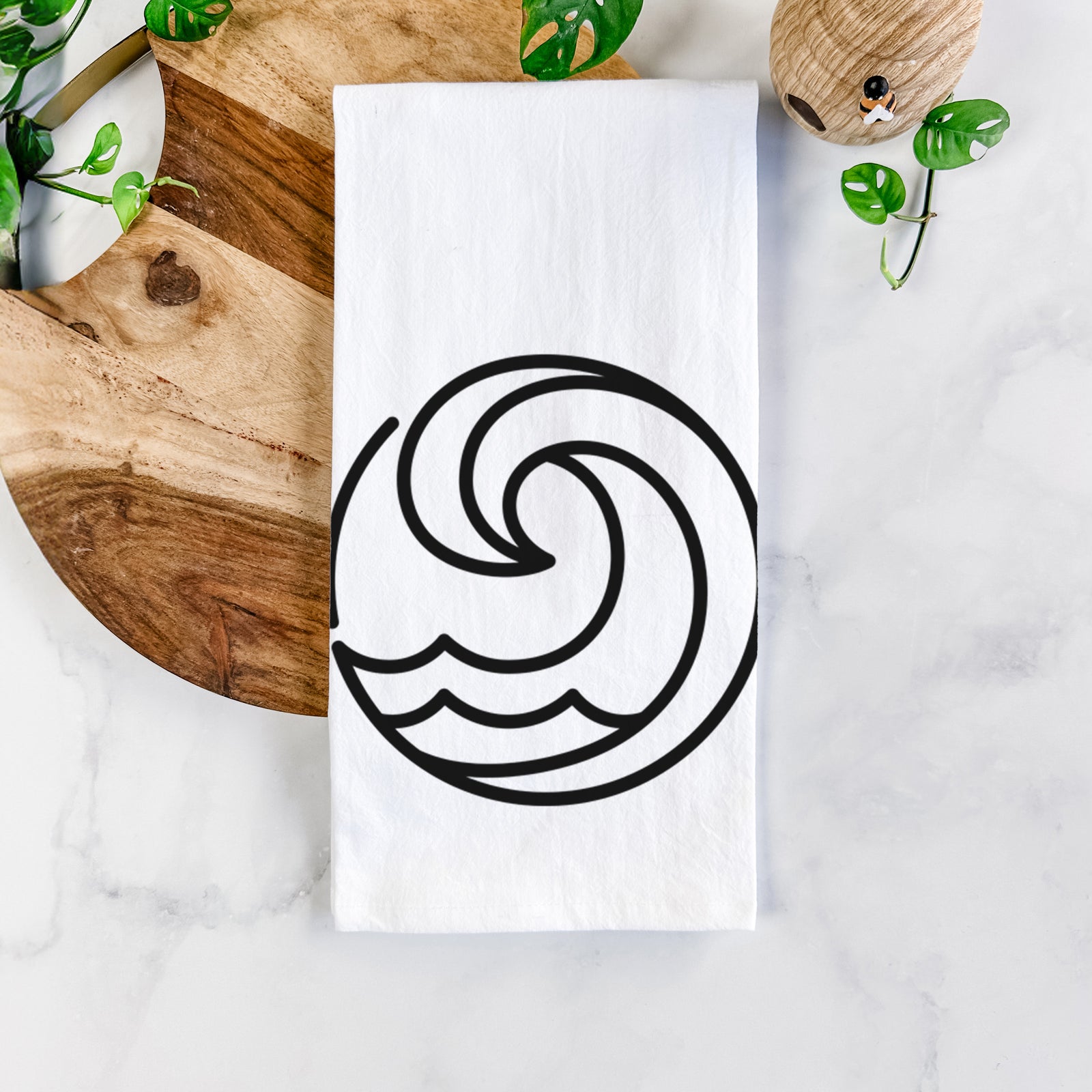 White cotton tea towel printed with a bold black line art circle of a curling ocean wave, on a marble counter beside a round wooden cutting board and greenery.