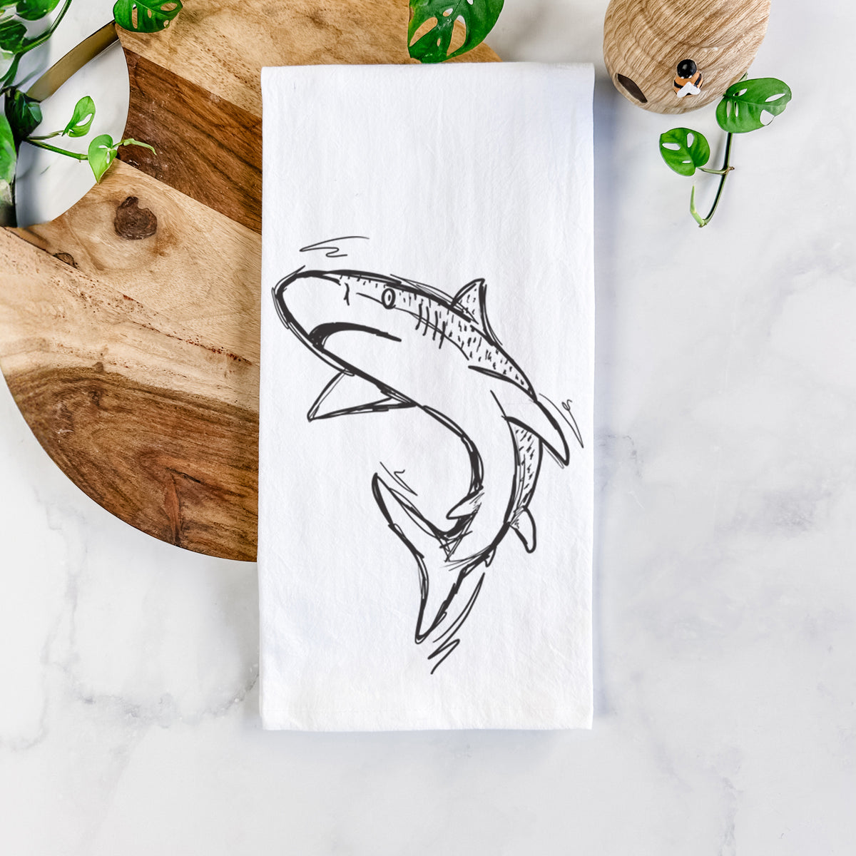 White cotton tea towel printed with a black ink illustration of a tiger shark swimming in a curve, on a marble counter beside a round wooden cutting board and greenery.