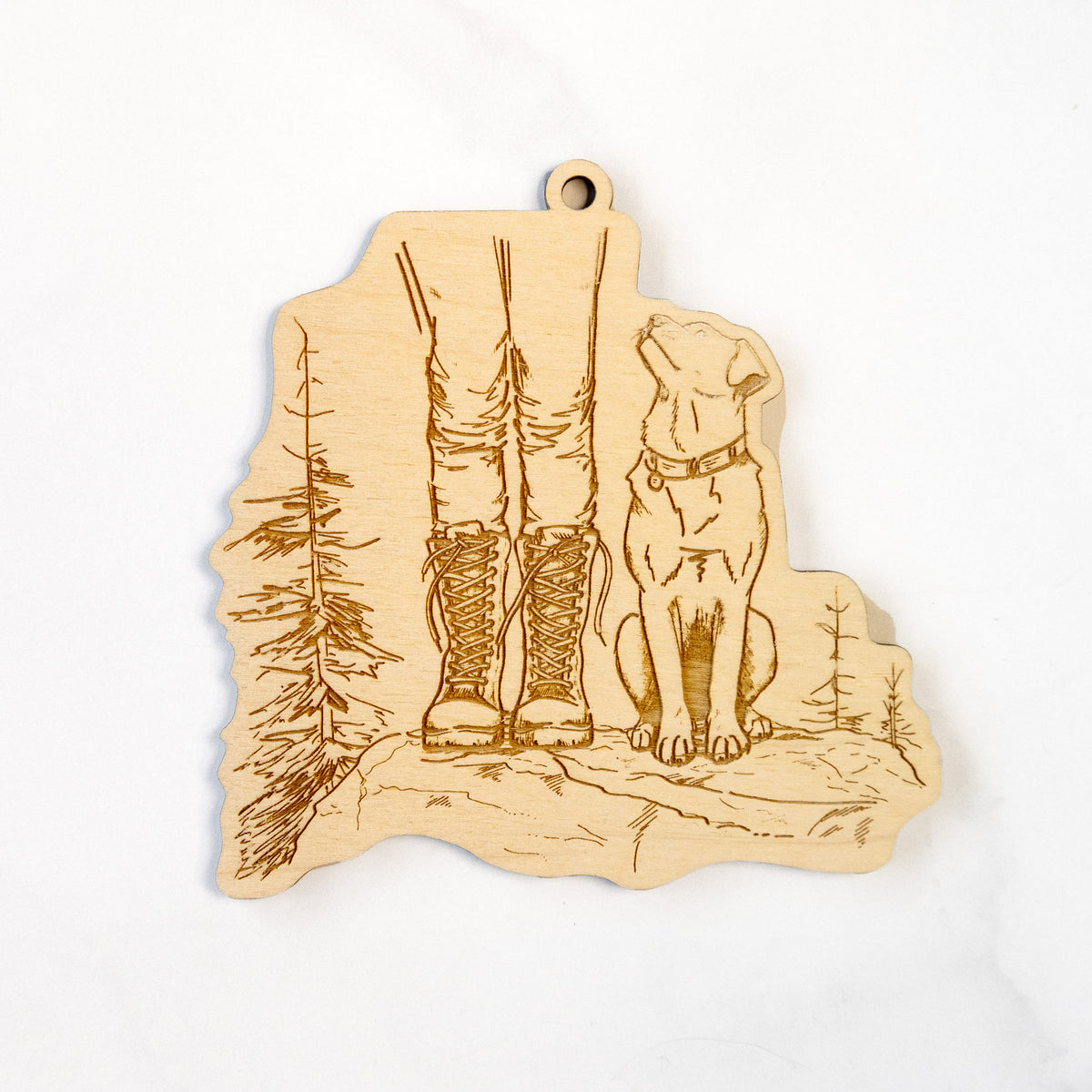 Wooden ornament featuring a engraving of a hiker in boots and a dog sitting side by side on a forest trail, seen from behind, on a marble surface.