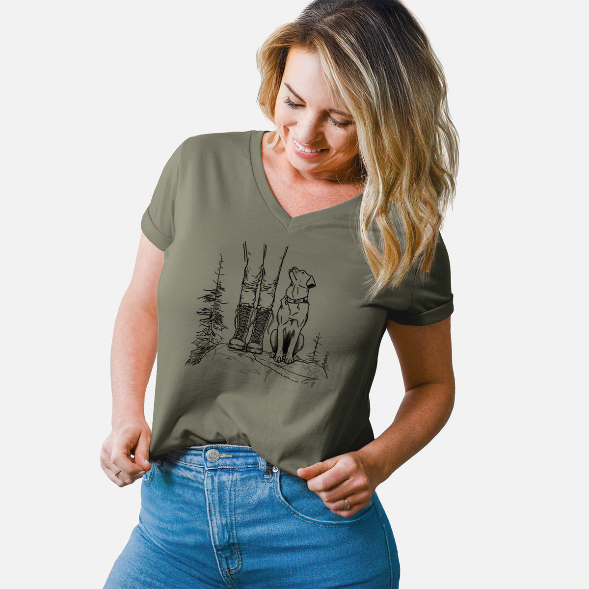 Woman wearing a military green women's relaxed fit cotton V-neck t-shirt printed with a black ink illustration of a hiker in boots and a dog sitting side by side on a forest trail, seen from behind.