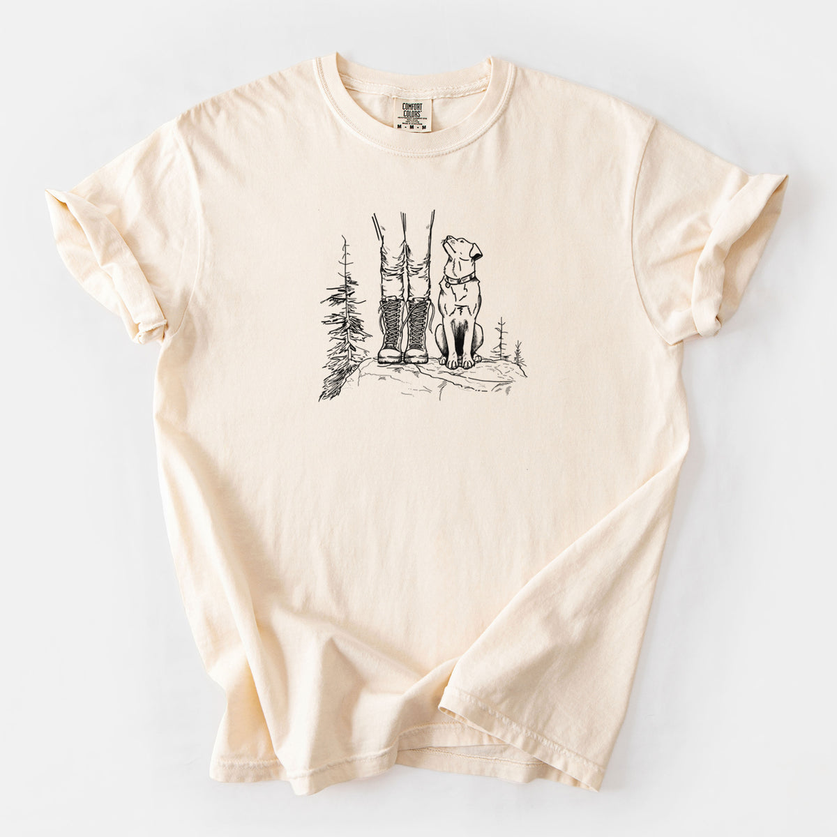 Ivory Comfort Colors t-shirt laid flat, printed with a black ink illustration of a hiker in boots and a dog sitting side by side on a forest trail, seen from behind.