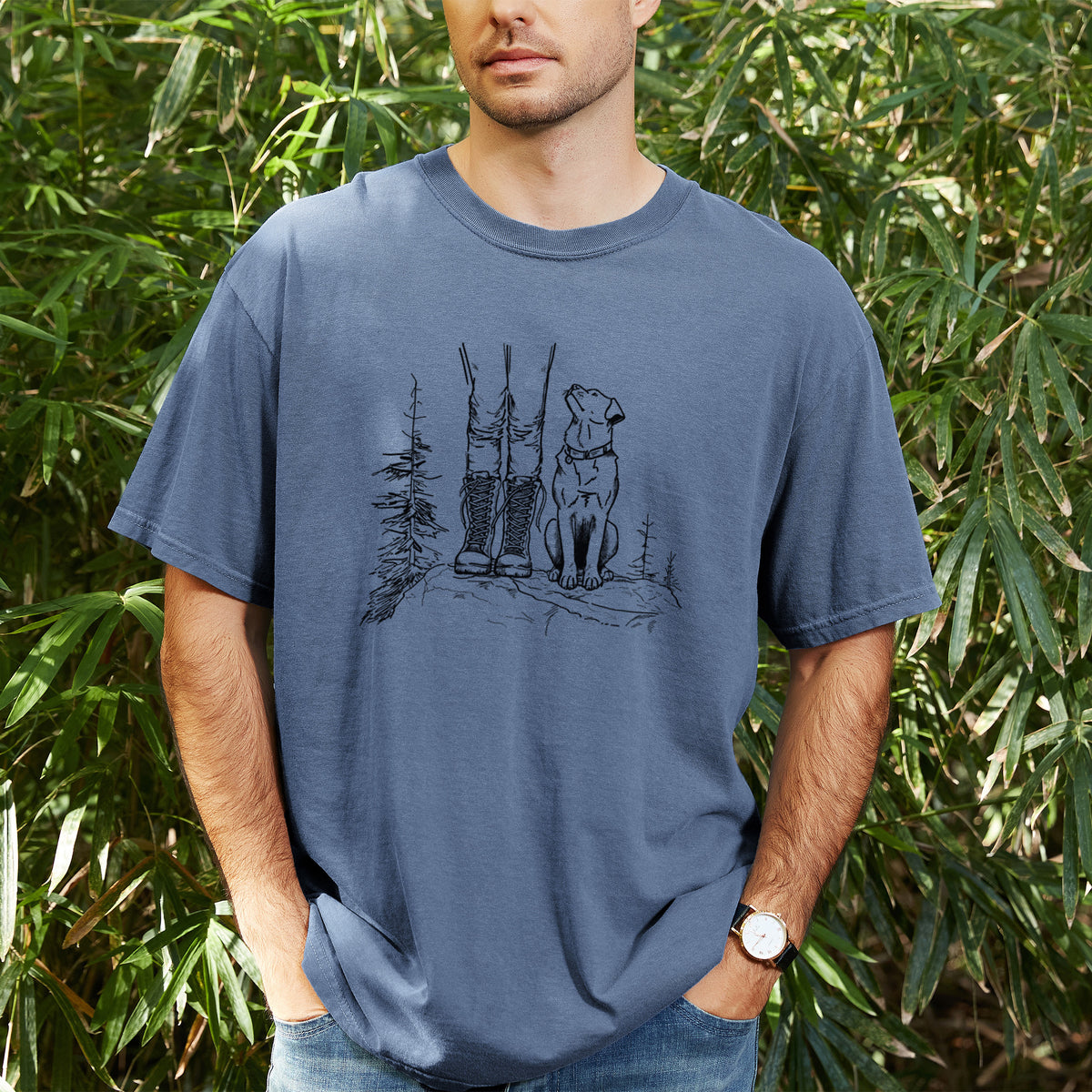 Man wearing a Comfort Colors t-shirt printed with a black ink illustration of a hiker in boots and a dog sitting side by side on a forest trail, seen from behind, outdoors.