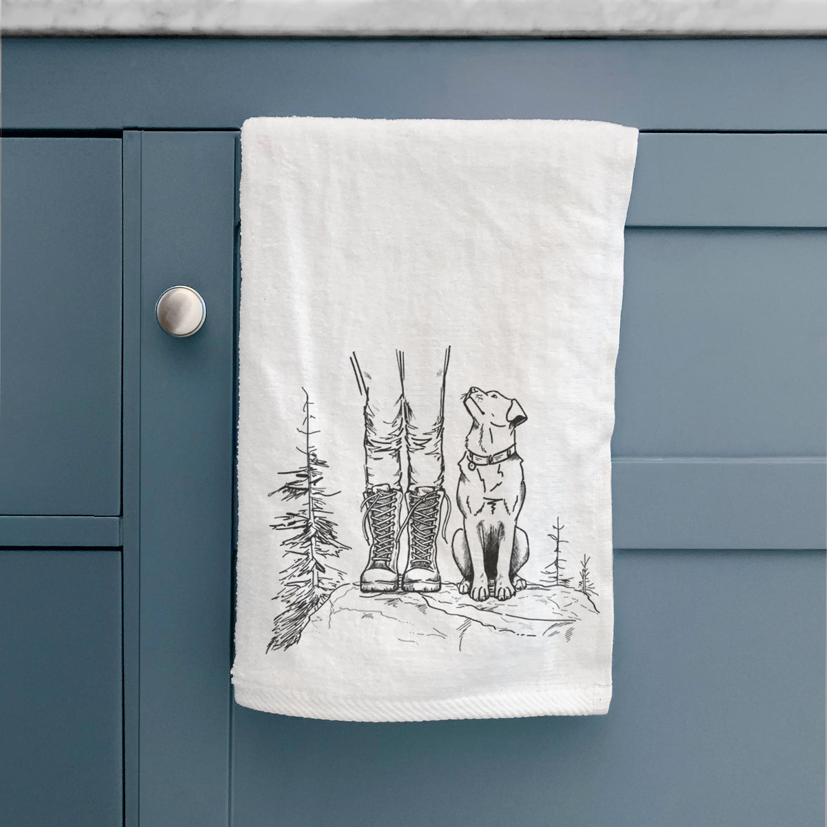 White hand towel printed with a black ink illustration of a hiker in boots and a dog sitting side by side on a forest trail, seen from behind, hanging on a cabinet door.