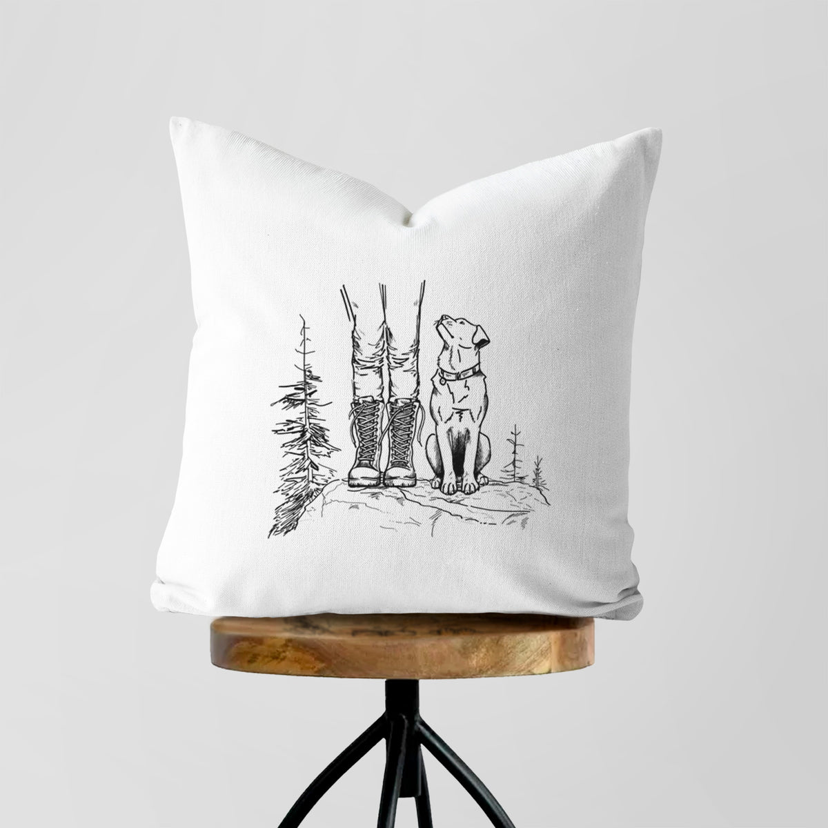White pillow cover printed with a black ink illustration of a hiker in boots and a dog sitting side by side on a forest trail, seen from behind, on a wooden stool.