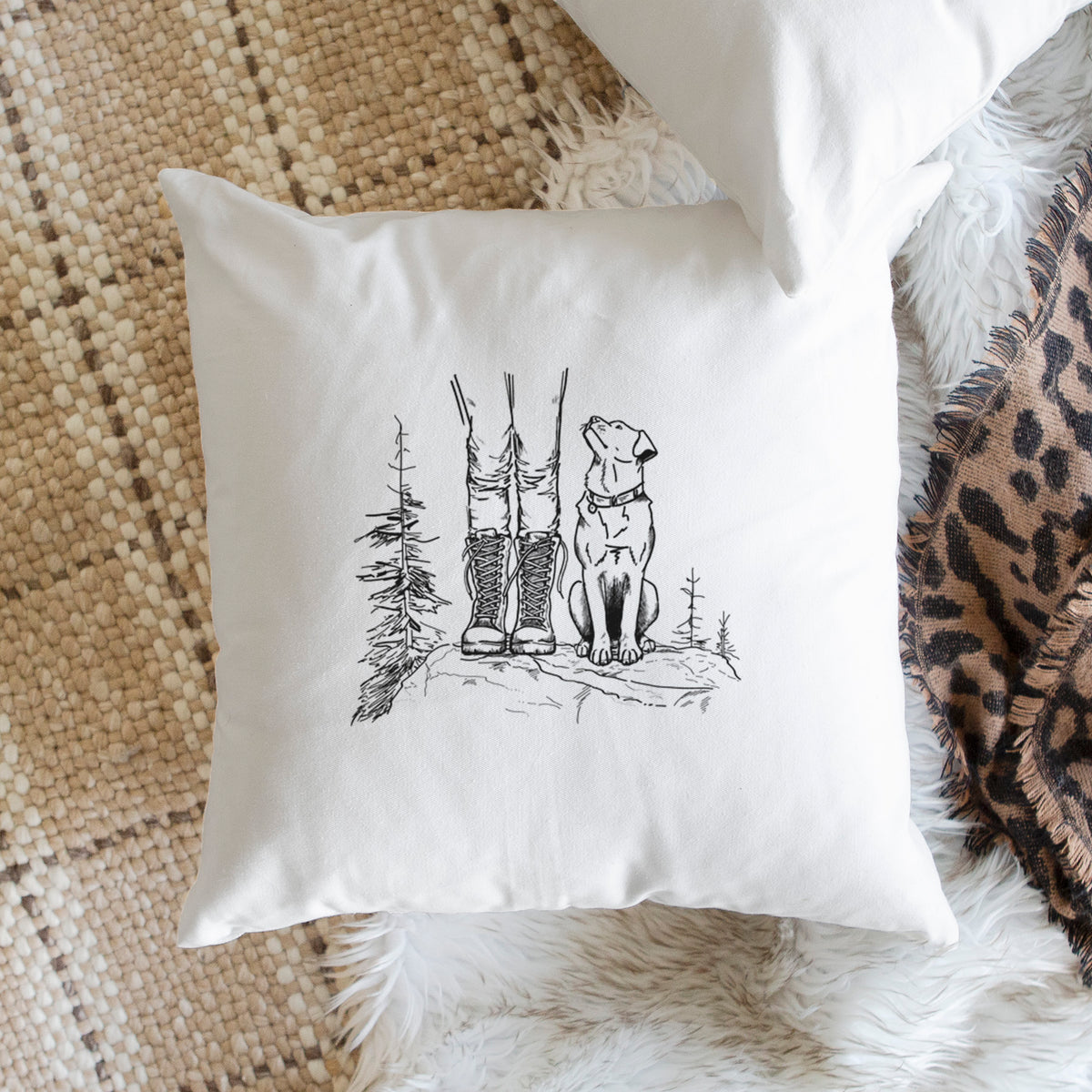 White pillow cover printed with a black ink illustration of a hiker in boots and a dog sitting side by side on a forest trail, seen from behind, on a woven chair.