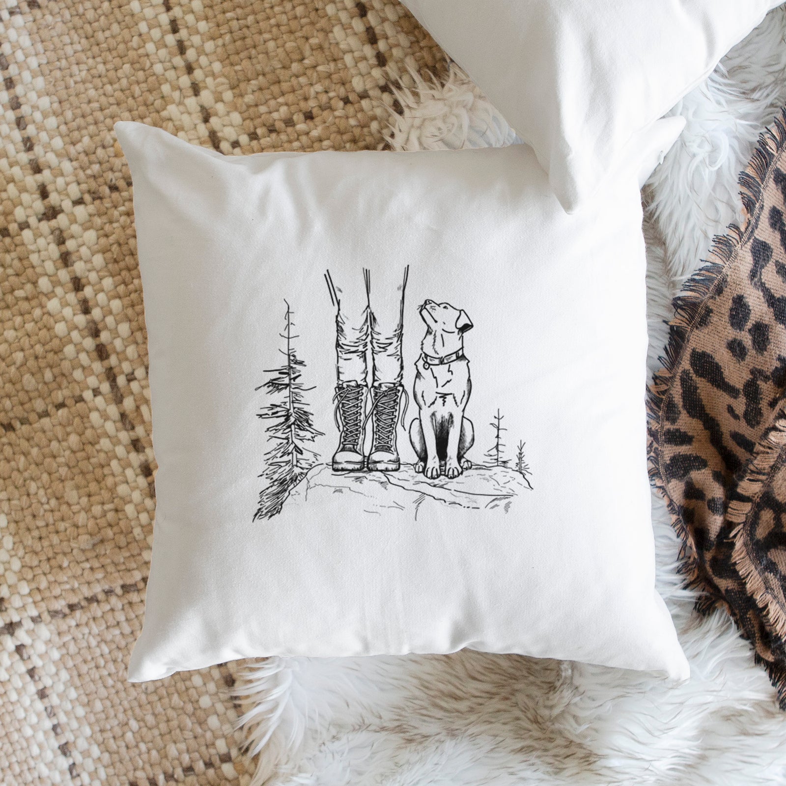 White pillow cover printed with a black ink illustration of a hiker in boots and a dog sitting side by side on a forest trail, seen from behind, on a bench beside plants.