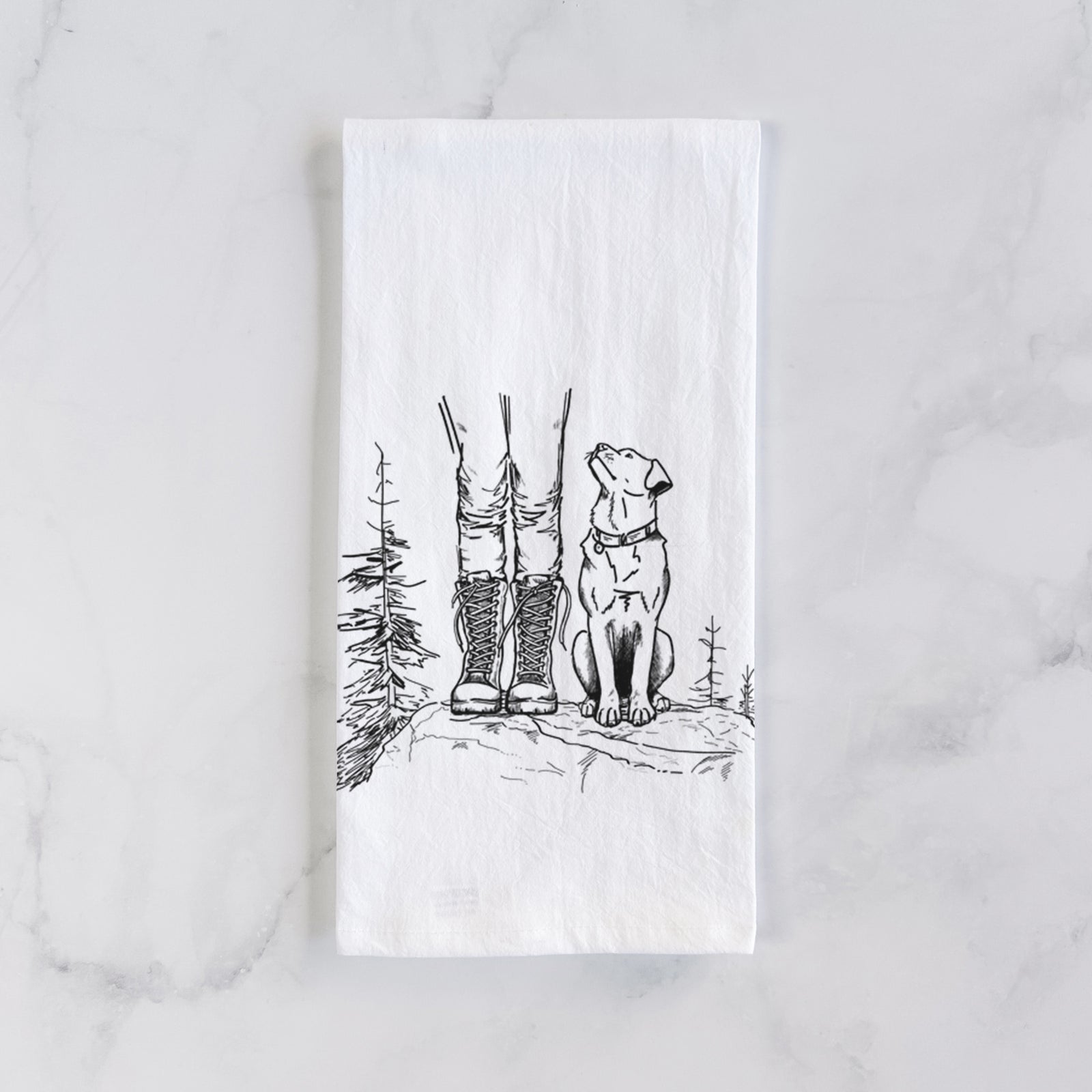 White cotton tea towel printed with a black ink illustration of a hiker in boots and a dog sitting side by side on a forest trail, seen from behind, on a marble counter beside a round wooden cutting board and greenery.