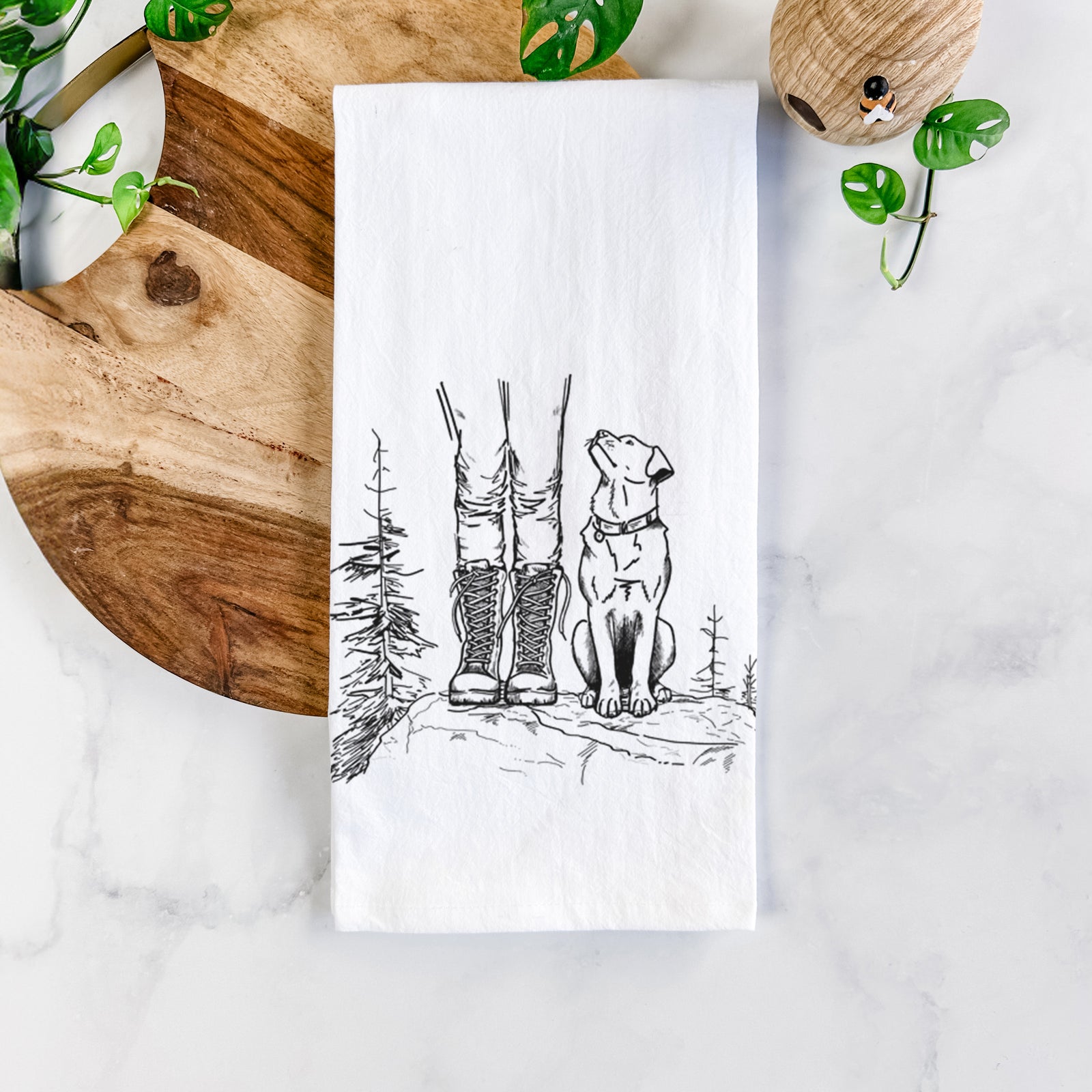 White cotton tea towel printed with a black ink illustration of a hiker in boots and a dog sitting side by side on a forest trail, seen from behind, on a marble counter beside a round wooden cutting board and greenery.
