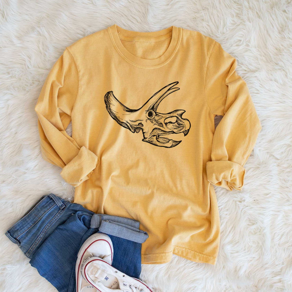 Golden wheat yellow Comfort Colors long sleeve shirt printed with a detailed black ink illustration of a Triceratops skull with its horns and frill, shown in a lifestyle photo.