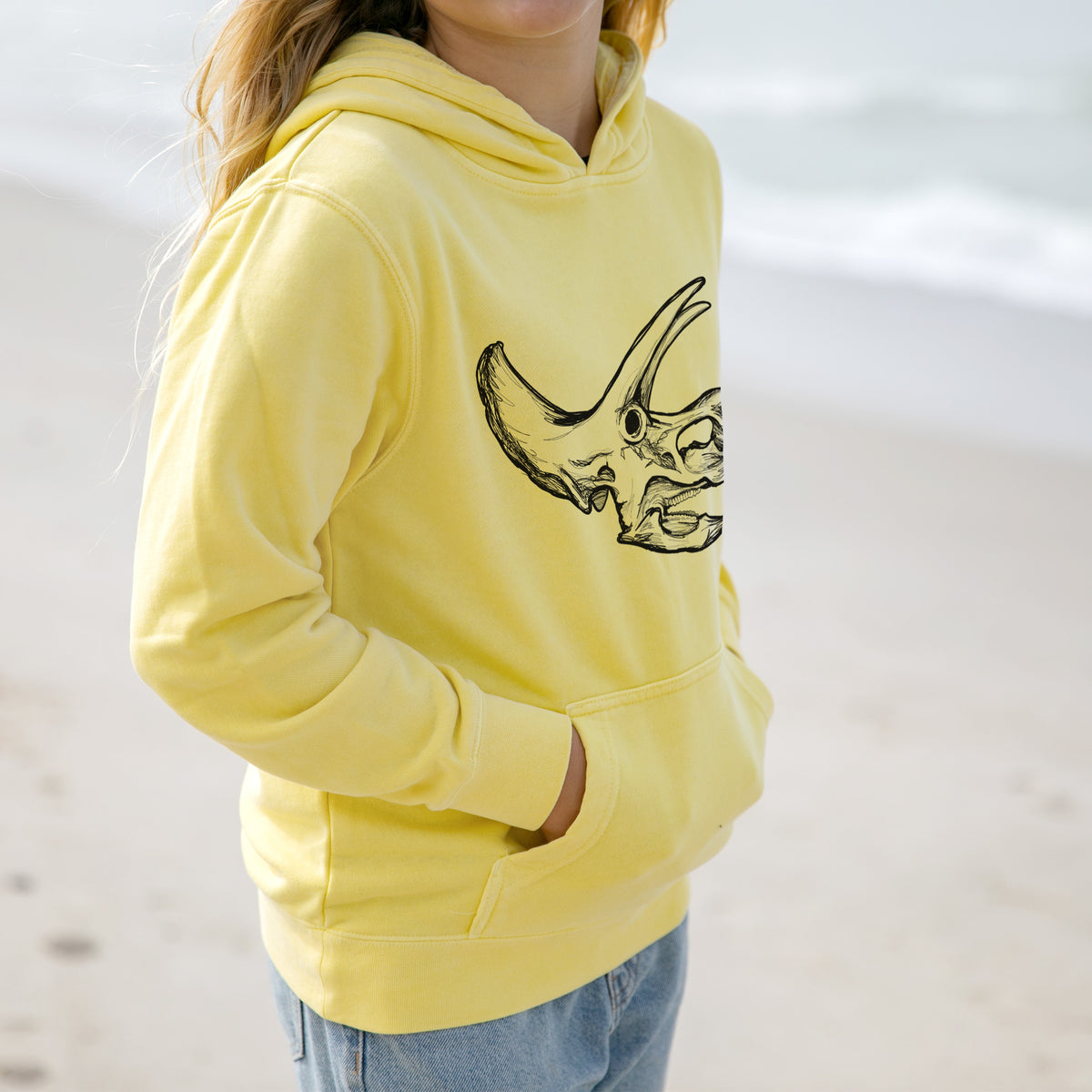 Girl wearing a youth pigment dyed hoodie printed with a detailed black ink illustration of a Triceratops skull with its horns and frill.