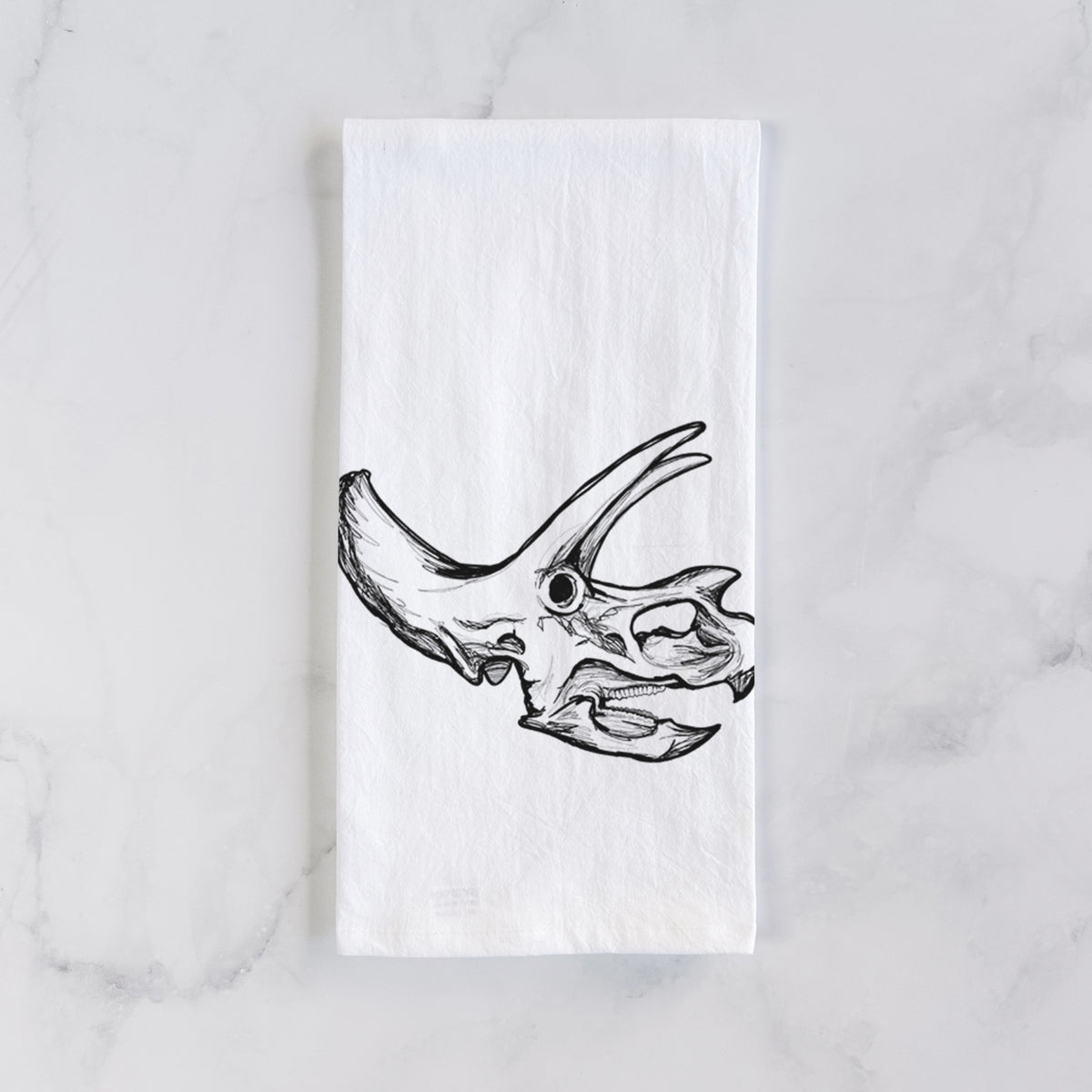 White cotton tea towel laid flat on marble, printed with a detailed black ink illustration of a Triceratops skull with its horns and frill.