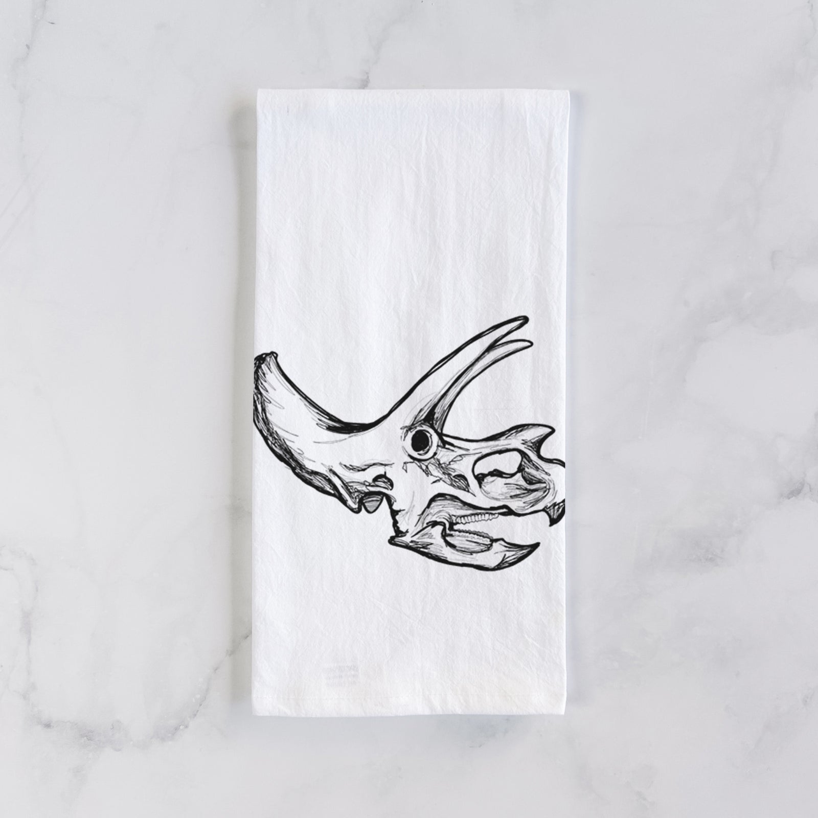 White cotton tea towel printed with a detailed black ink illustration of a Triceratops skull with its horns and frill, on a marble counter beside a round wooden cutting board and greenery.
