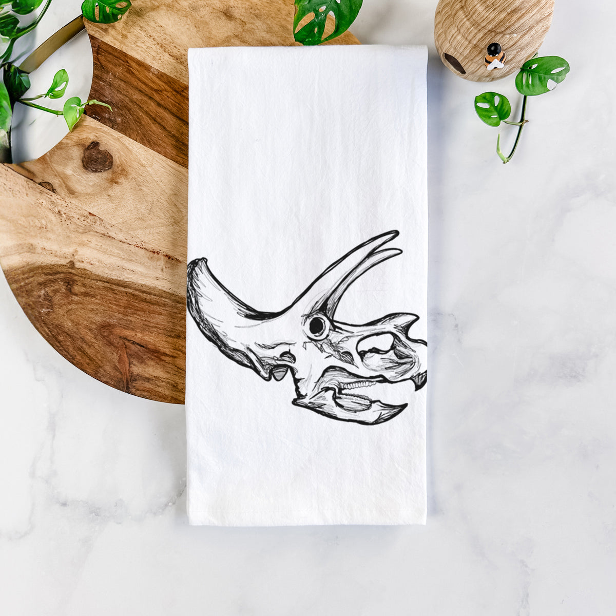 White cotton tea towel printed with a detailed black ink illustration of a Triceratops skull with its horns and frill, on a marble counter beside a round wooden cutting board and greenery.