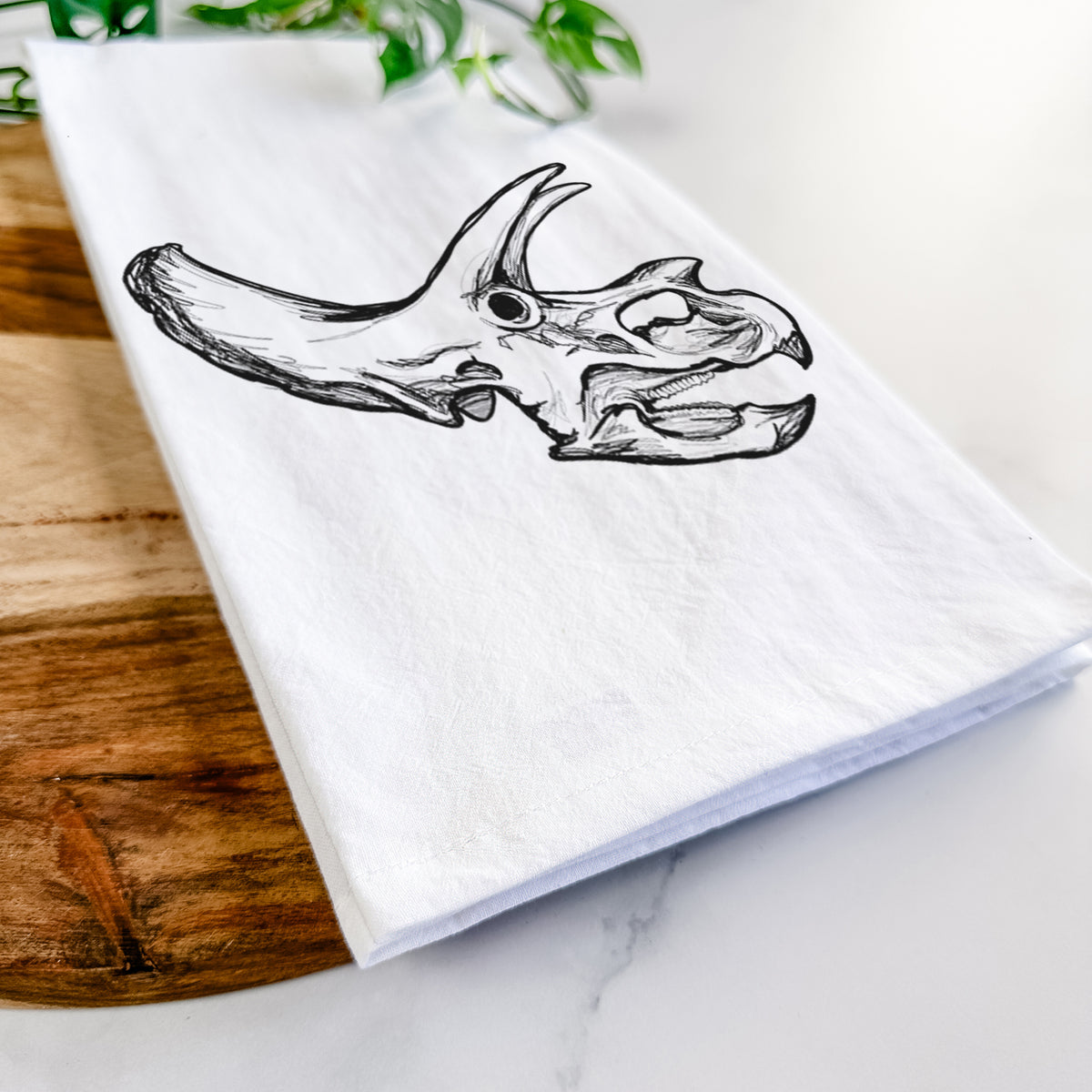 Close-up of a folded white tea towel on a wooden board, showing a detailed black ink illustration of a Triceratops skull with its horns and frill.