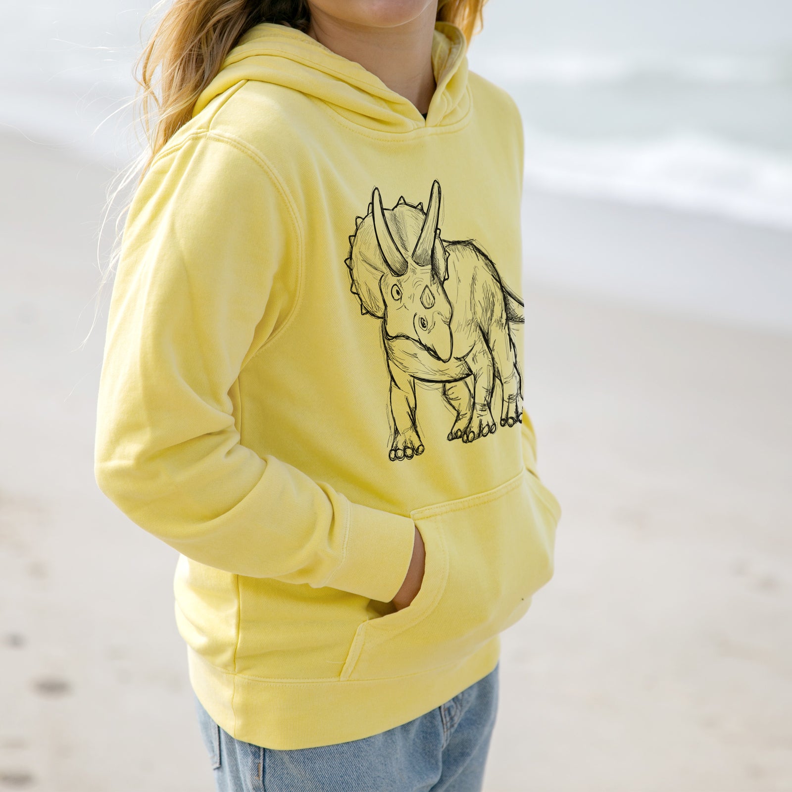 Boy wearing a youth pigment dyed hoodie printed with a black ink illustration of a Triceratops walking.