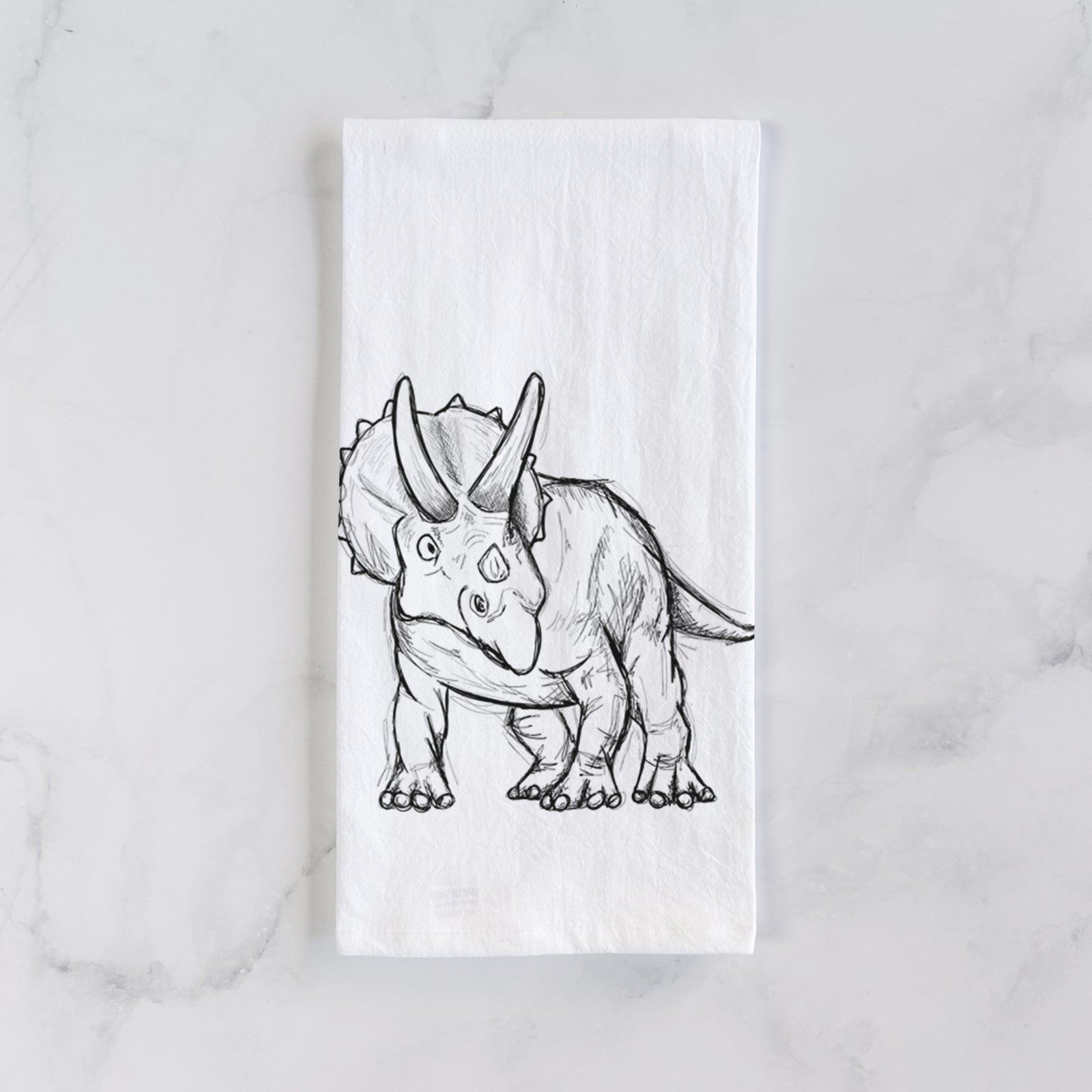 White cotton tea towel printed with a black ink illustration of a Triceratops walking, on a marble counter beside a round wooden cutting board and greenery.