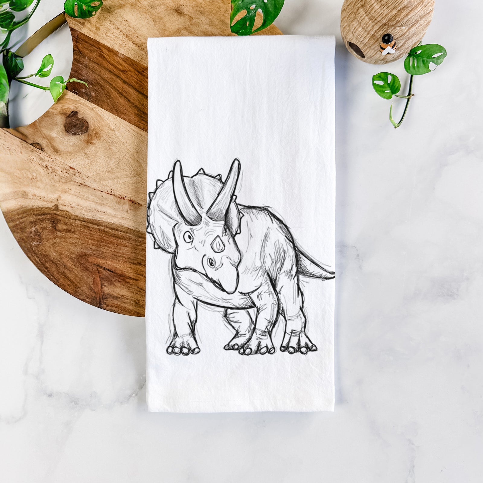 White cotton tea towel printed with a black ink illustration of a Triceratops walking, on a marble counter beside a round wooden cutting board and greenery.