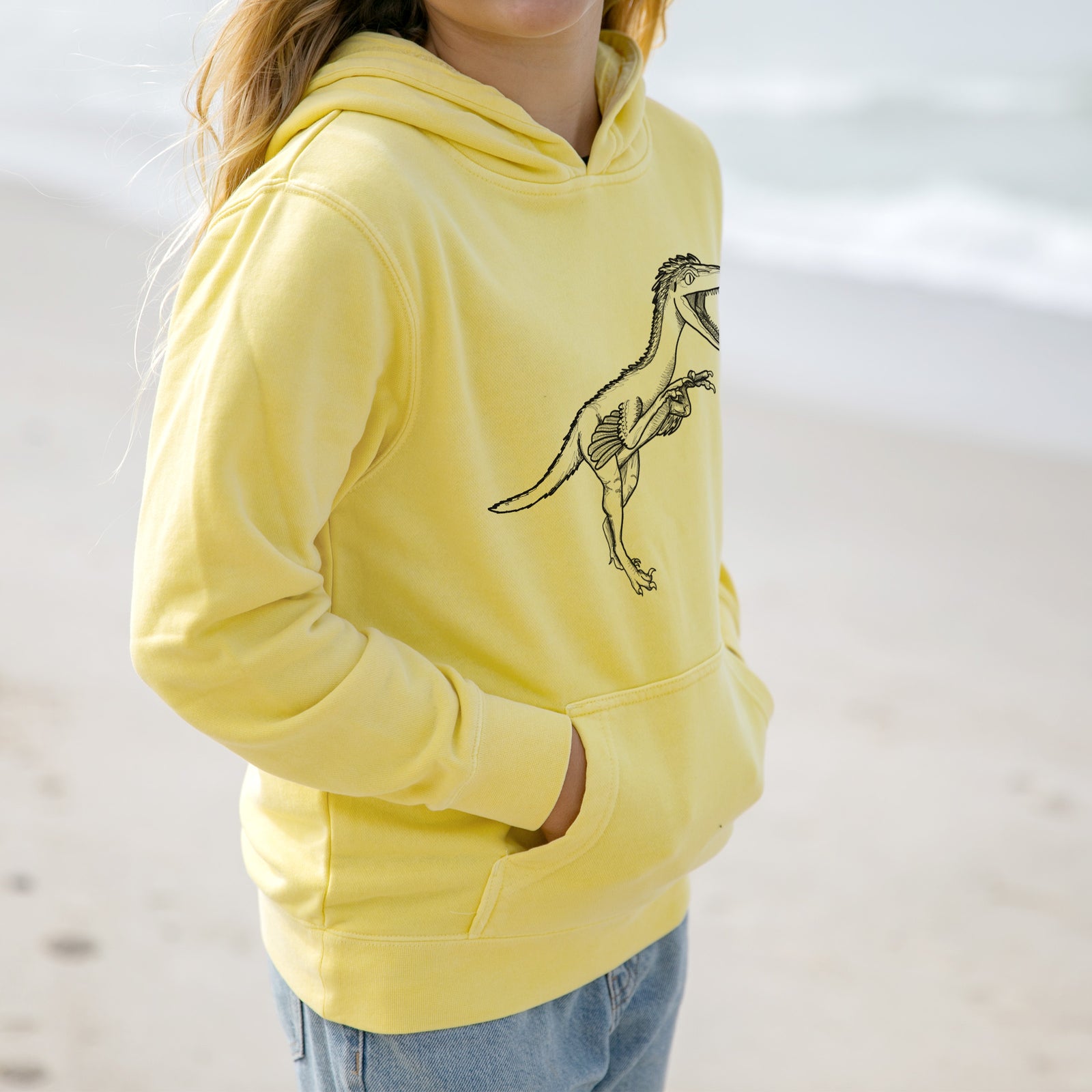 Boy wearing a youth pigment dyed hoodie printed with a black ink illustration of a Troodon dinosaur running.