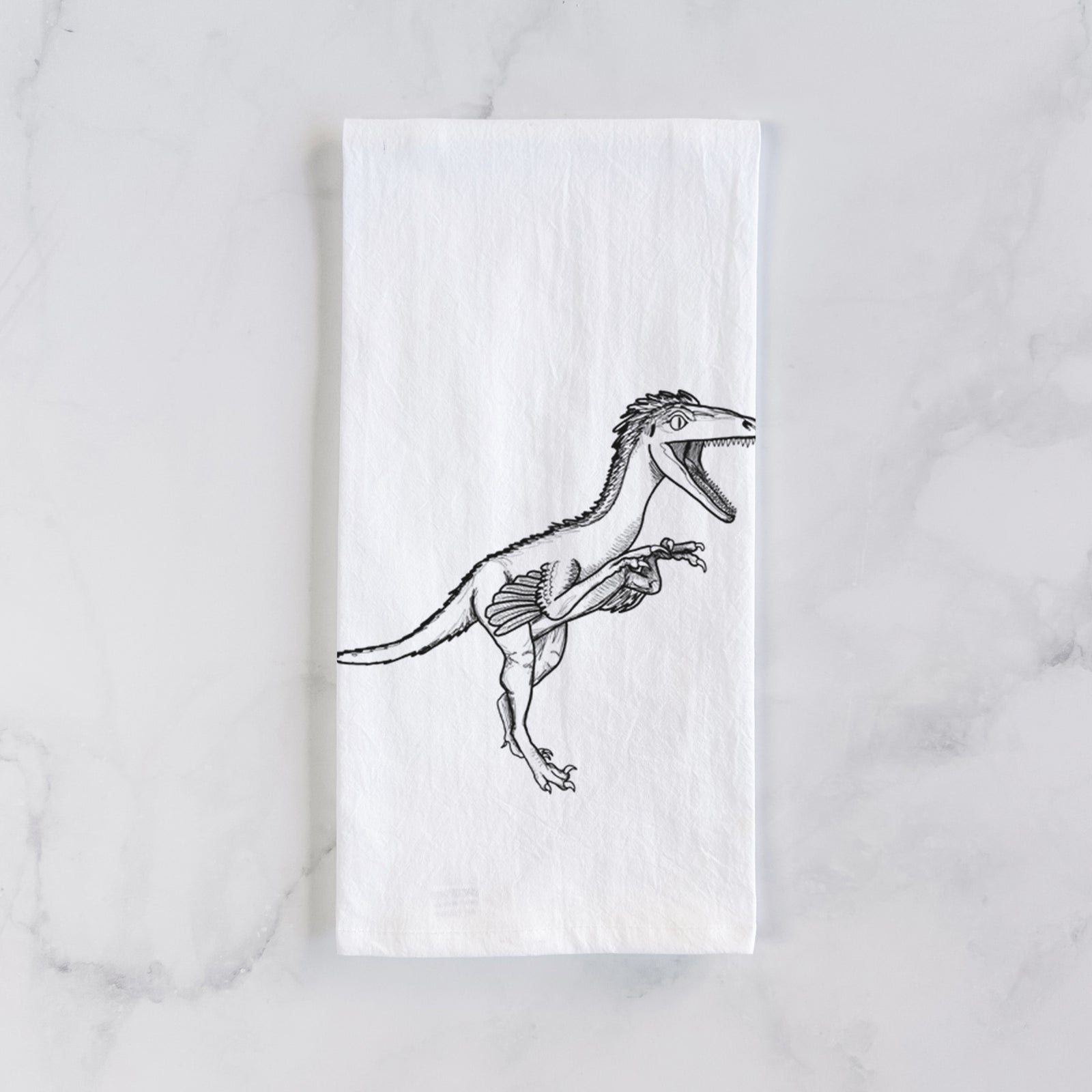 White cotton tea towel printed with a black ink illustration of a Troodon dinosaur running, on a marble counter beside a round wooden cutting board and greenery.