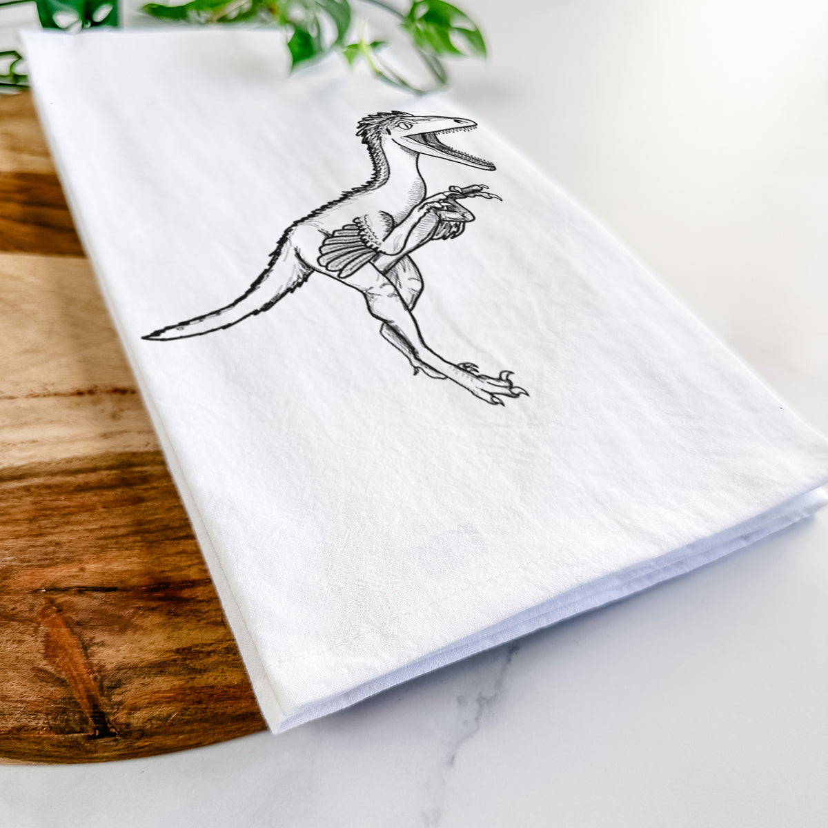 Close-up of a folded white tea towel on a wooden board, showing a black ink illustration of a Troodon dinosaur running.