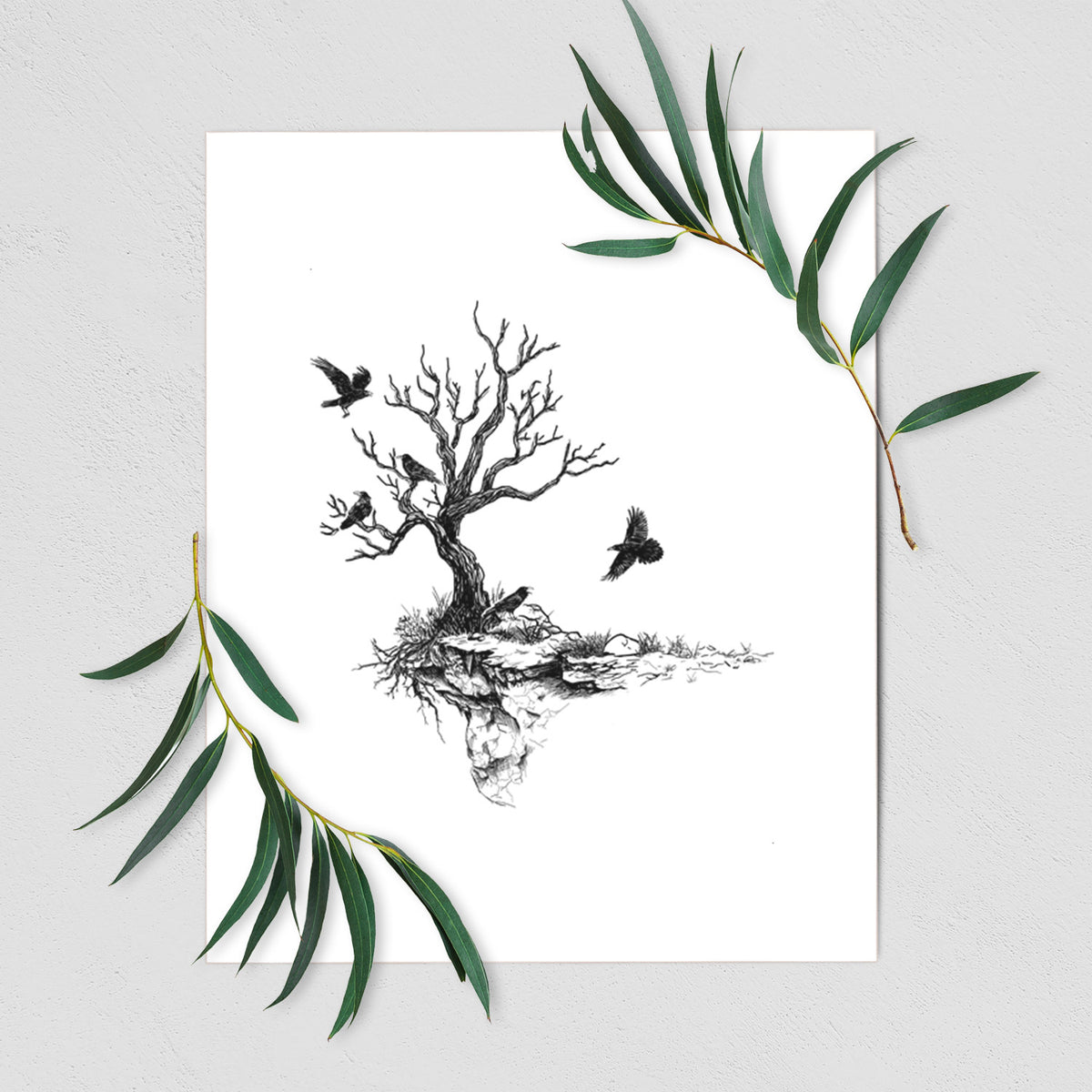 Art print of a black ink illustration of a bare twisted tree on a small island with ravens flying around it, shown with eucalyptus leaves.