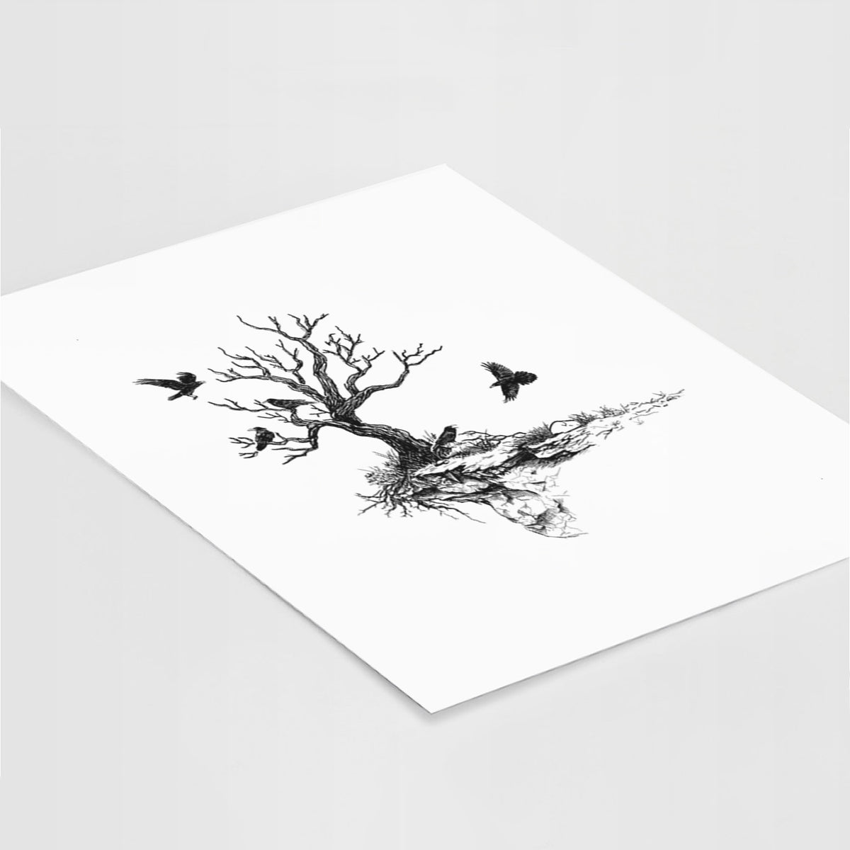 Art print of a black ink illustration of a bare twisted tree on a small island with ravens flying around it, lying at an angle on a white surface.