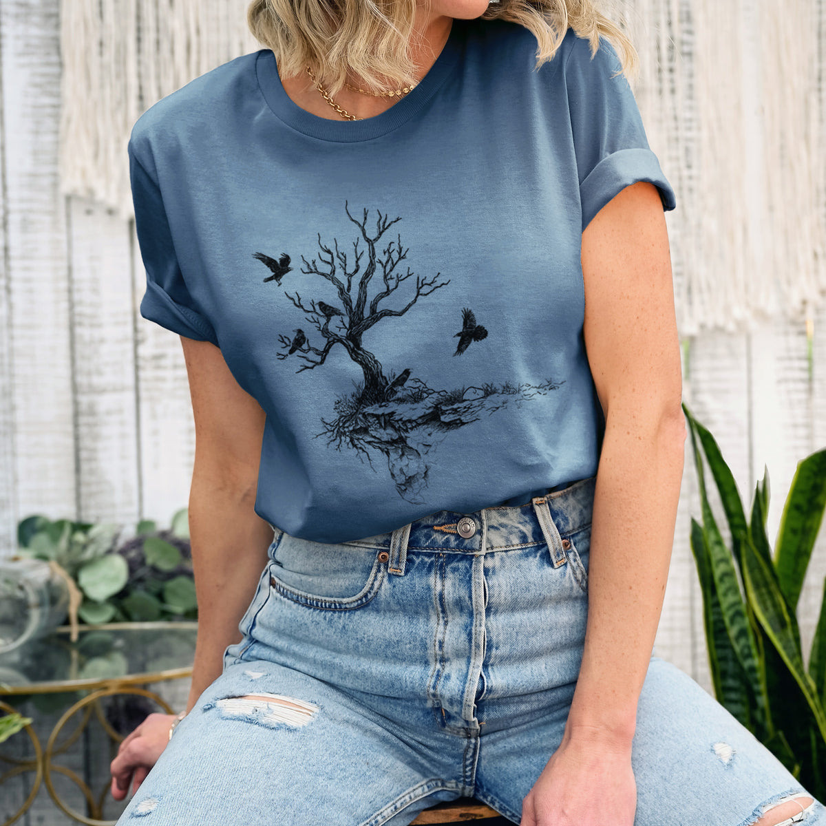 Woman wearing a steel blue lightweight cotton t-shirt printed with a black ink illustration of a bare twisted tree on a small island with ravens flying around it.