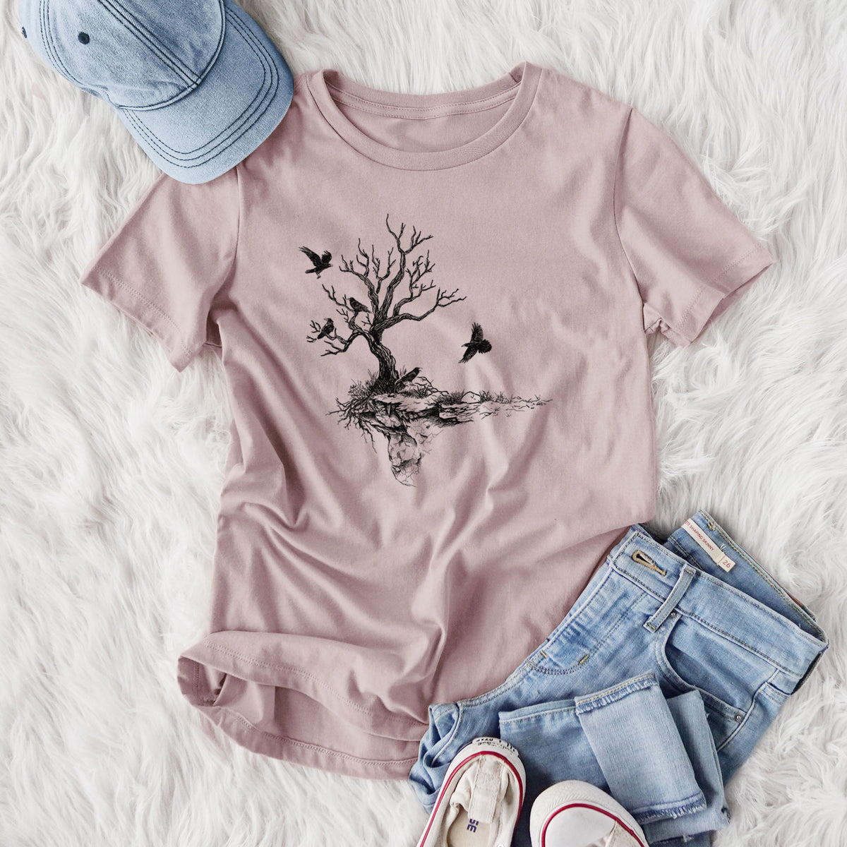 Women's relaxed fit cotton t-shirt printed with a black ink illustration of a bare twisted tree on a small island with ravens flying around it, styled flat on a white faux fur rug with jeans.