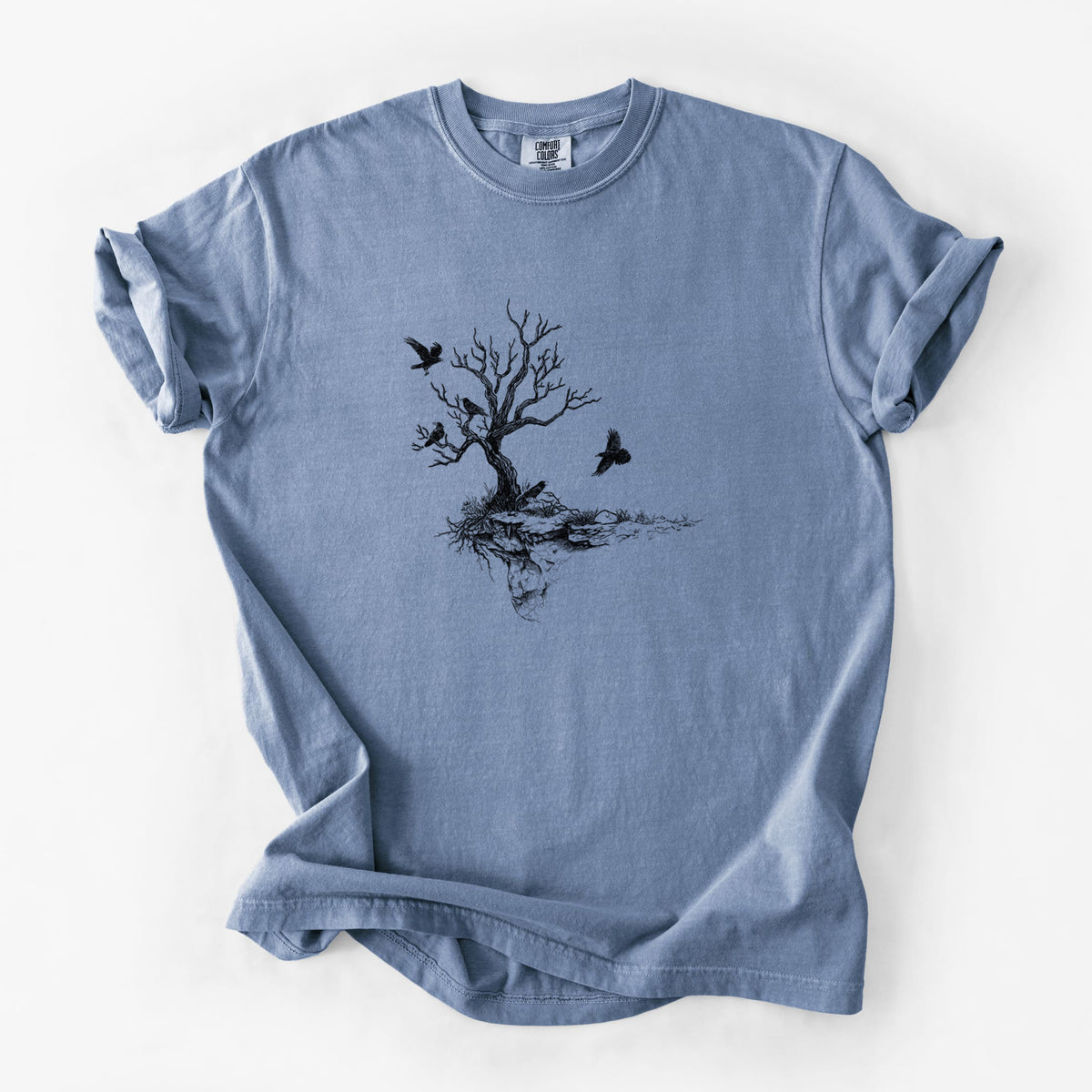 Blue jean Comfort Colors t-shirt laid flat, printed with a black ink illustration of a bare twisted tree on a small island with ravens flying around it.