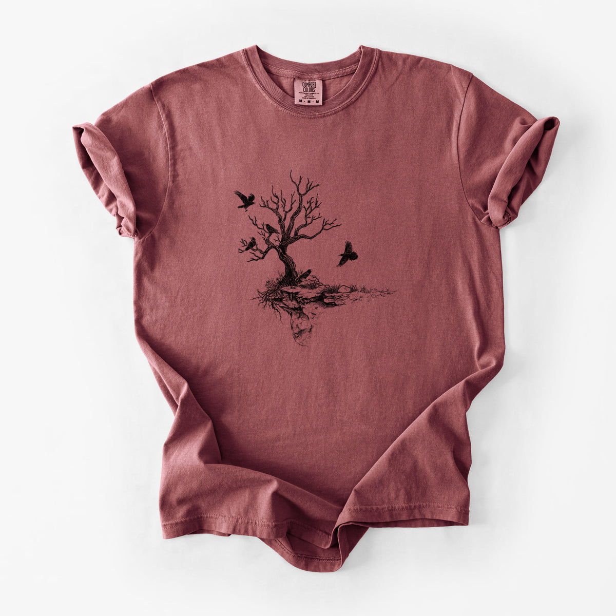 Brick red Comfort Colors t-shirt laid flat, printed with a black ink illustration of a bare twisted tree on a small island with ravens flying around it.