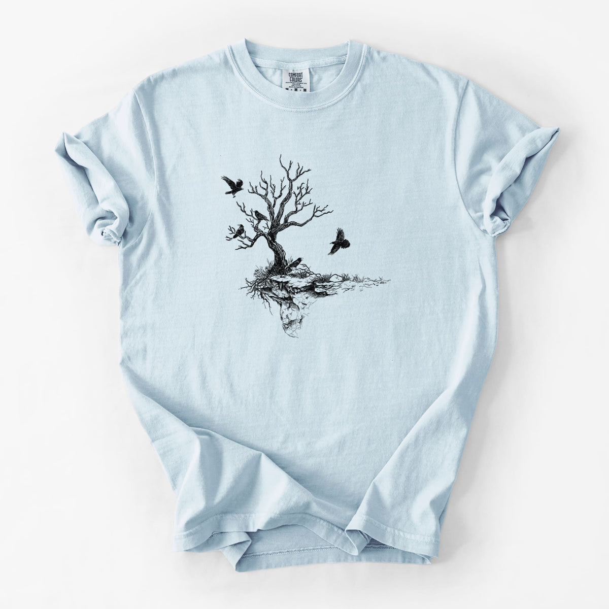 Chambray blue Comfort Colors t-shirt laid flat, printed with a black ink illustration of a bare twisted tree on a small island with ravens flying around it.