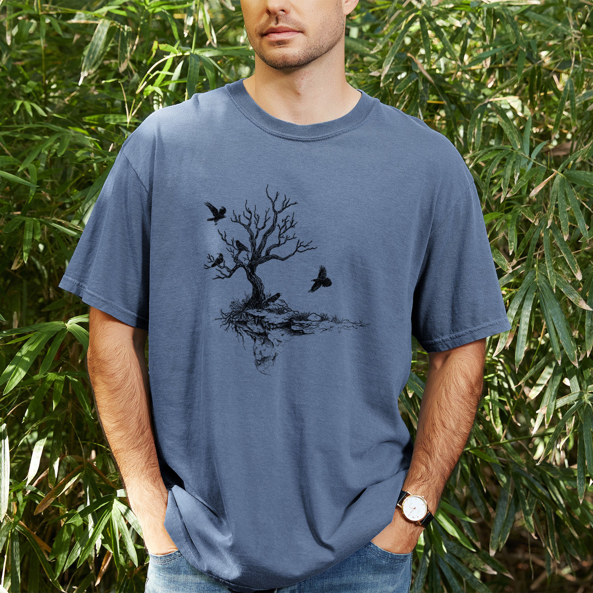 Man wearing a Comfort Colors t-shirt printed with a black ink illustration of a bare twisted tree on a small island with ravens flying around it, outdoors.