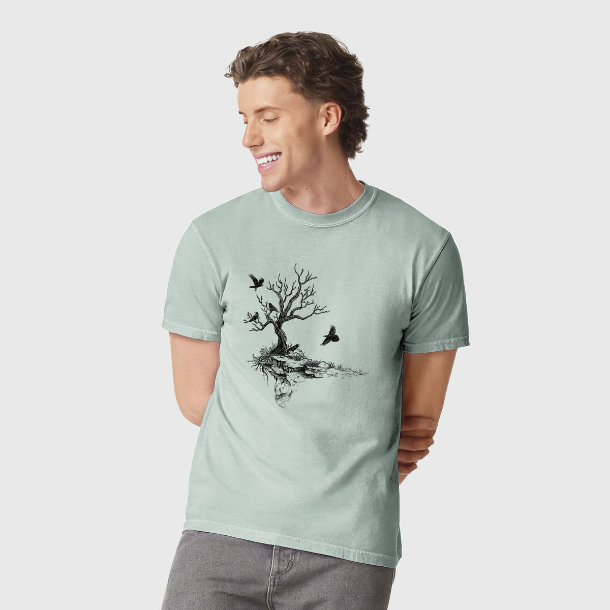 Man wearing a Comfort Colors t-shirt printed with a black ink illustration of a bare twisted tree on a small island with ravens flying around it.