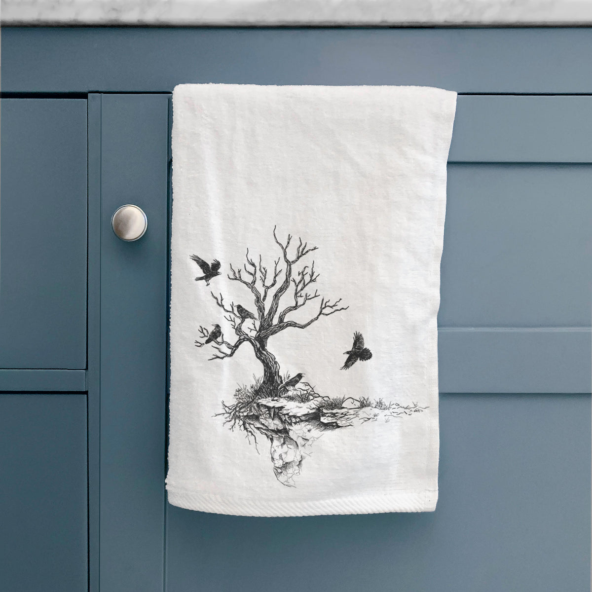 White hand towel printed with a black ink illustration of a bare twisted tree on a small island with ravens flying around it, hanging on a cabinet door.