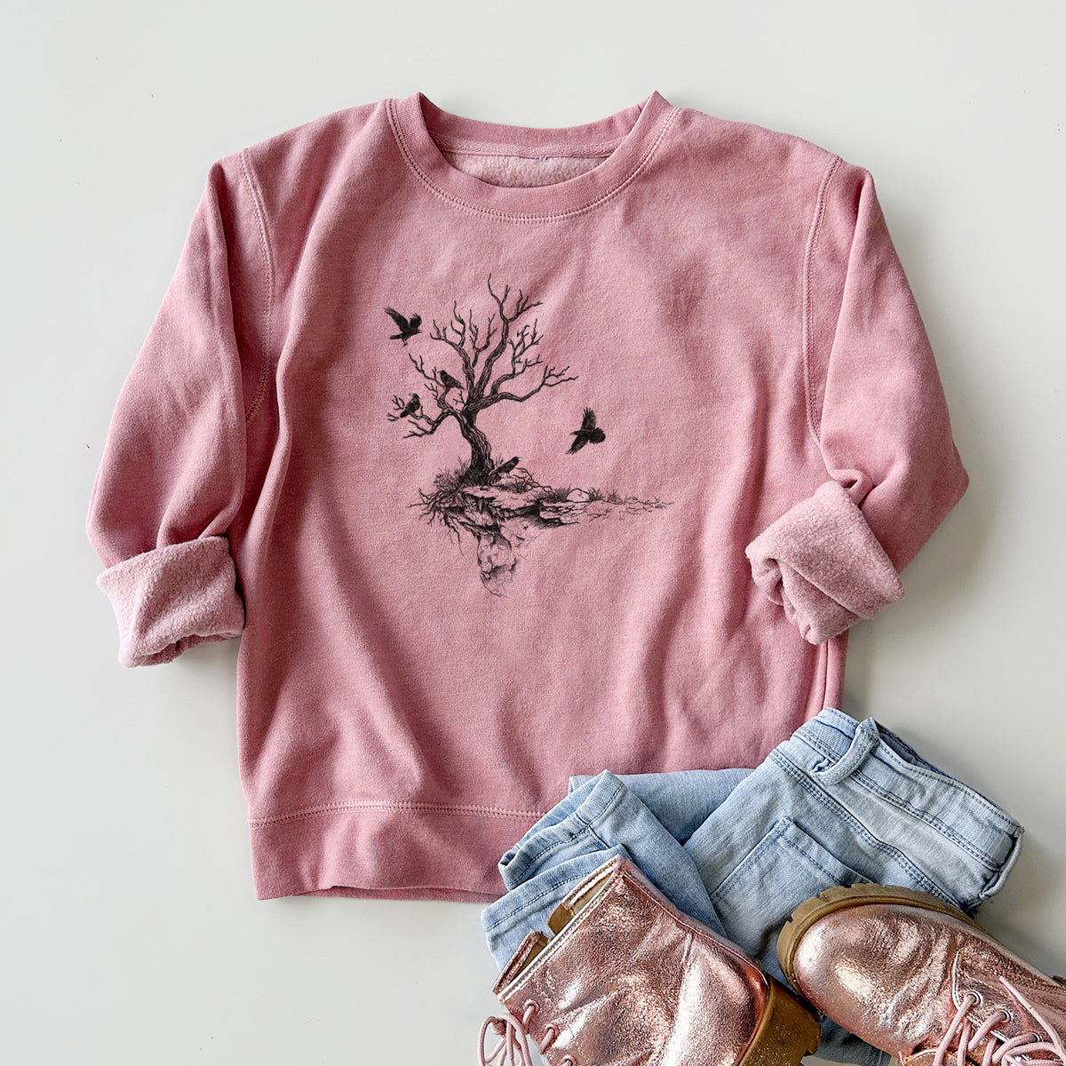 Youth crewneck sweatshirt printed with a black ink illustration of a bare twisted tree on a small island with ravens flying around it, styled flat with jeans and boots.