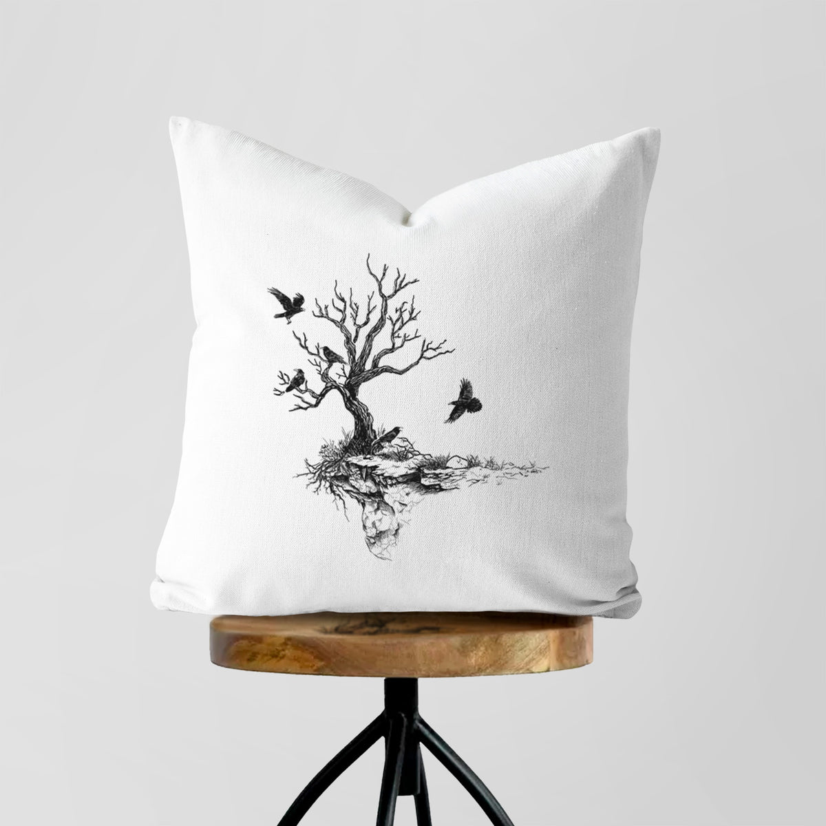 White pillow cover printed with a black ink illustration of a bare twisted tree on a small island with ravens flying around it, on a wooden stool.