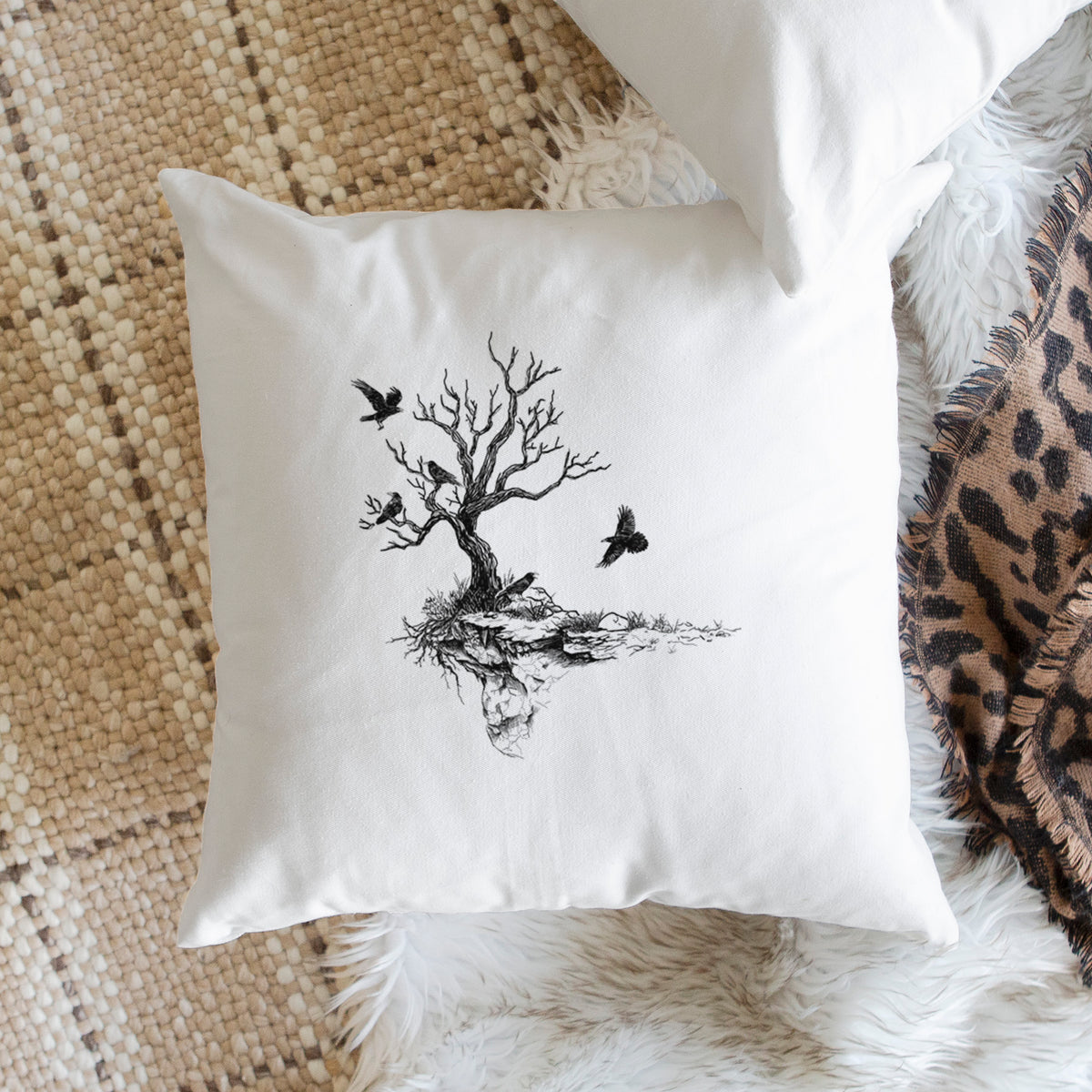 White pillow cover printed with a black ink illustration of a bare twisted tree on a small island with ravens flying around it, on a woven chair.