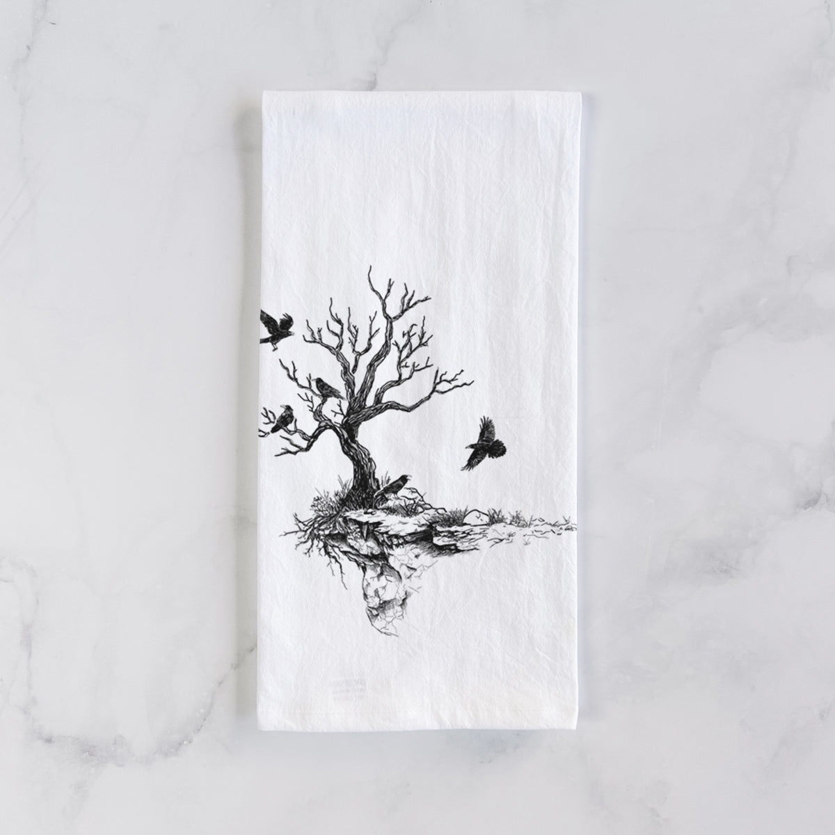White cotton tea towel laid flat on marble, printed with a black ink illustration of a bare twisted tree on a small island with ravens flying around it.