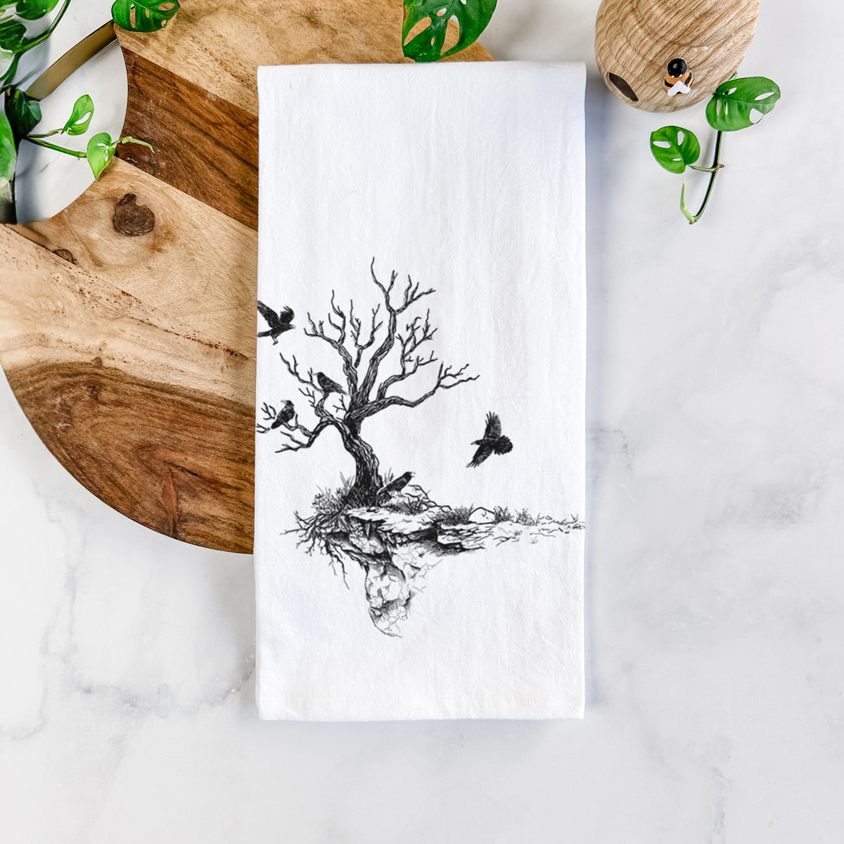 White cotton tea towel printed with a black ink illustration of a bare twisted tree on a small island with ravens flying around it, on a marble counter beside a round wooden cutting board and greenery.
