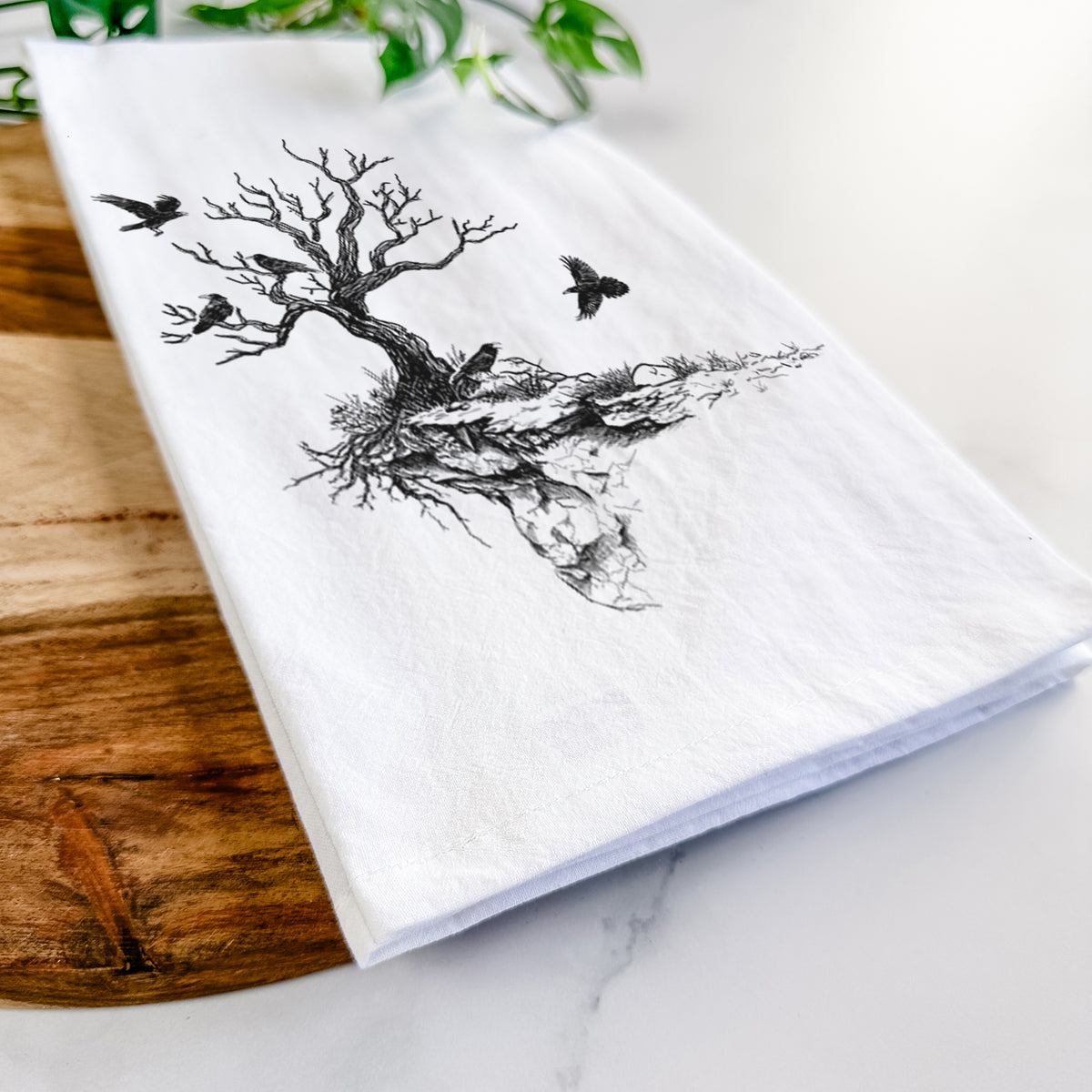 Close-up of a folded white tea towel on a wooden board, showing a black ink illustration of a bare twisted tree on a small island with ravens flying around it.