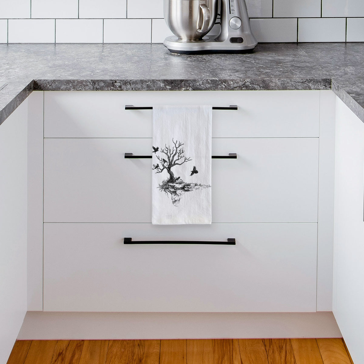 White tea towel printed with a black ink illustration of a bare twisted tree on a small island with ravens flying around it, hanging from a drawer handle in a bright white kitchen.