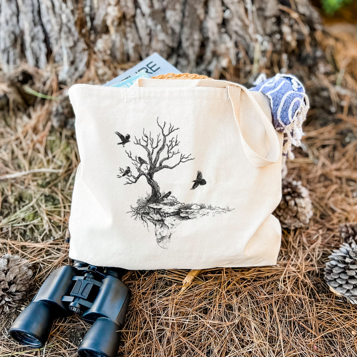 Canvas tote bag printed with a black ink illustration of a bare twisted tree on a small island with ravens flying around it, resting on dry grass beside a pair of binoculars.